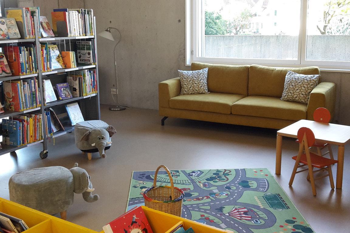 In einer Bibliothek steht ein gelbes Sofa vor einem Fenster. Davor liegt ein Spielteppich und vor einem Büchergestell stehen zwei Hocker in Elefantenform.