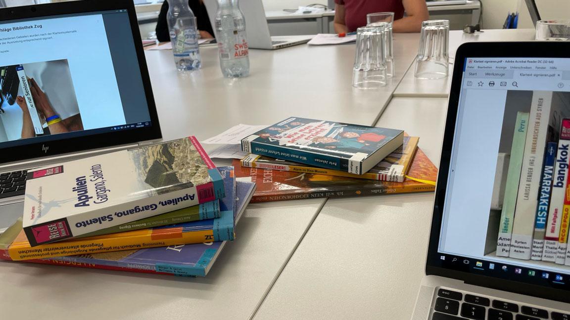 Auf einem Tisch, zwischen zwei Laptops, liegen Bücher die mit Klartextsystematik beschriftet sind.