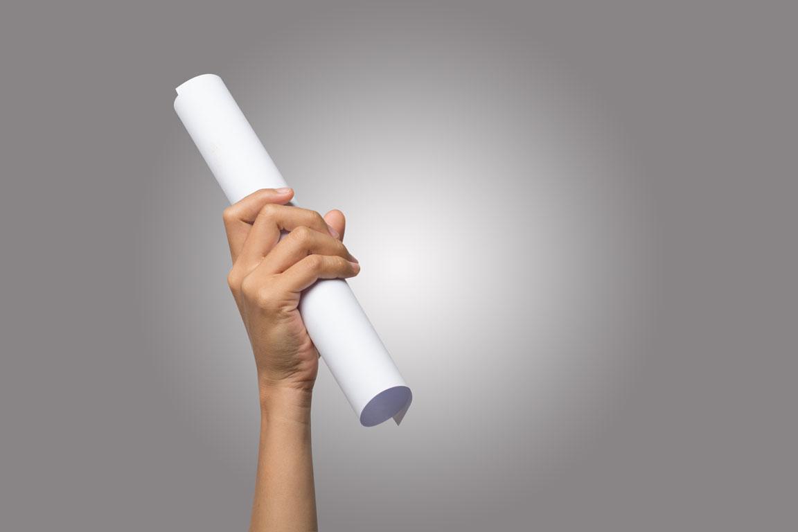 Eine Frauenhand hält vor einem grauen Hintergrund ein gerolltes Blatt Papier in die Höhe.