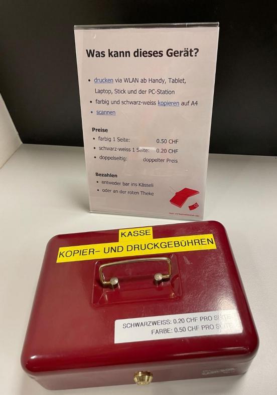 Auf einem Tisch steht eine rote Geldkassette vor einer Preisliste eines Druckers.