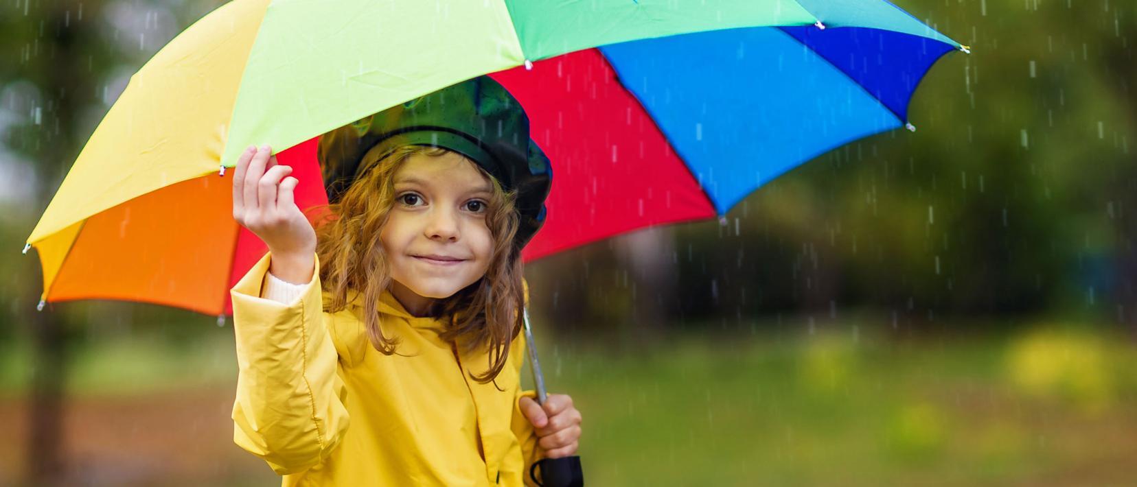 Kleines, lächelndes Mädchen steht mit mehrfarbigem Regenschirm in einem regnerischen Park.