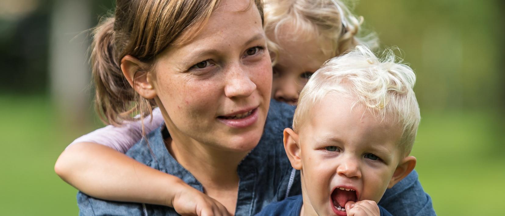 Mutter mit zwei Kleinkindern im Freien. Die Mutter hält eines der Kinder im Arm, das andere befindet sich im Hintergrund.