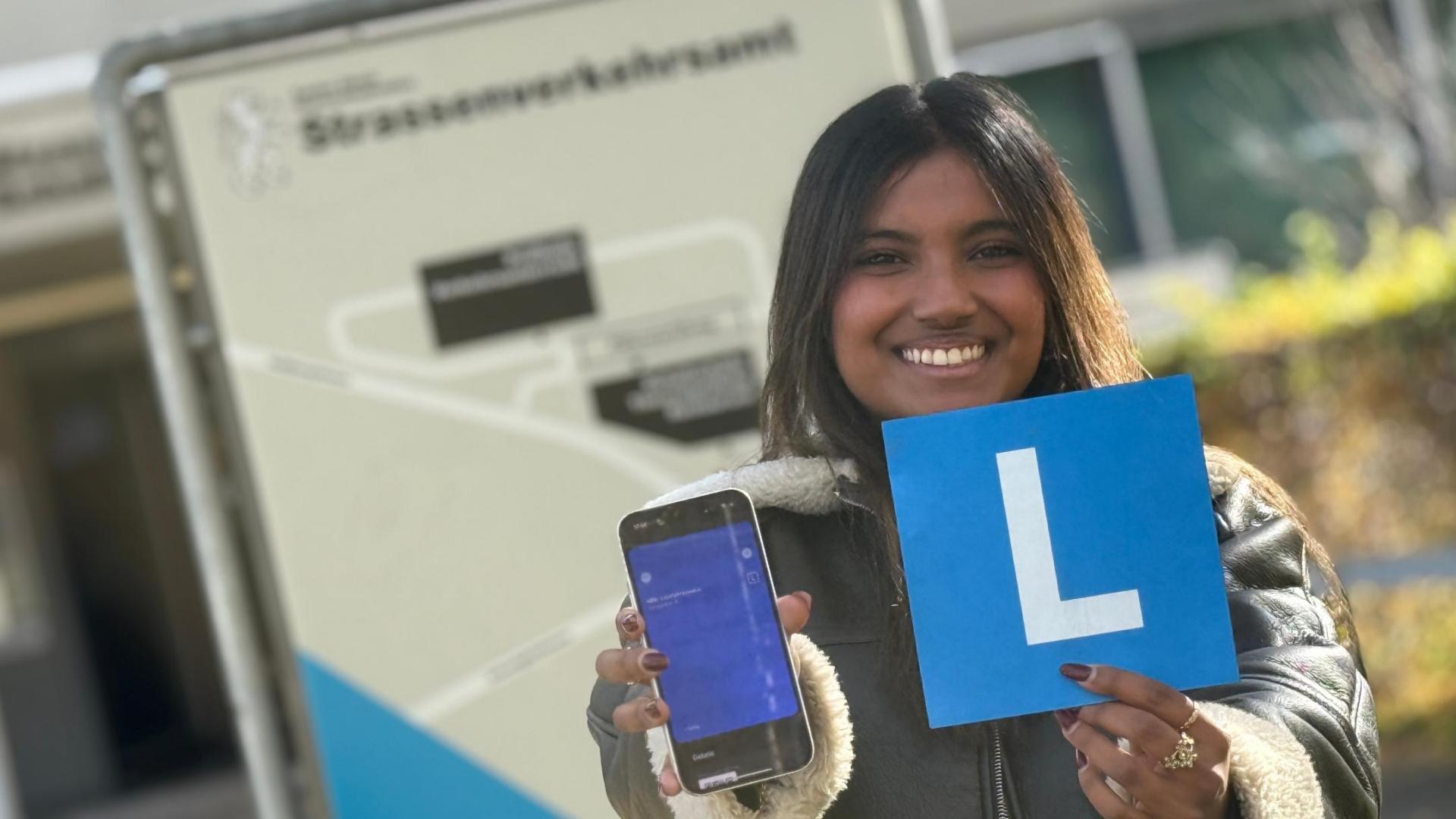 Eine Lernfahrerin steht vor dem Strassenverkehrsamt und hält ihr Smartphone mit dem elektronischen Lernfahrausweis sowie das blaue L-Schild in die Kamera.