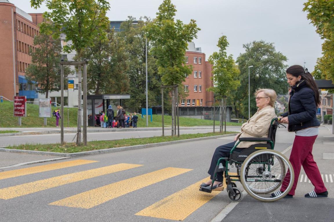 Eine Rollstuhlfahrerin wird von einer Pflegerin über einen Fussgängerstreifen gestossen.