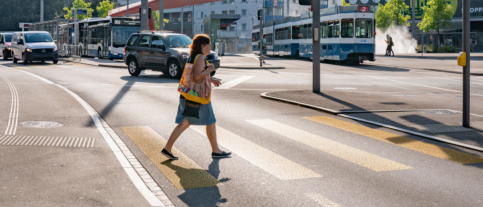 Eine Fussgängerin überquert einen signalisierten Fussgängerstreifen, im Hintergrund fahren Autos, ein Bus und eine Tram