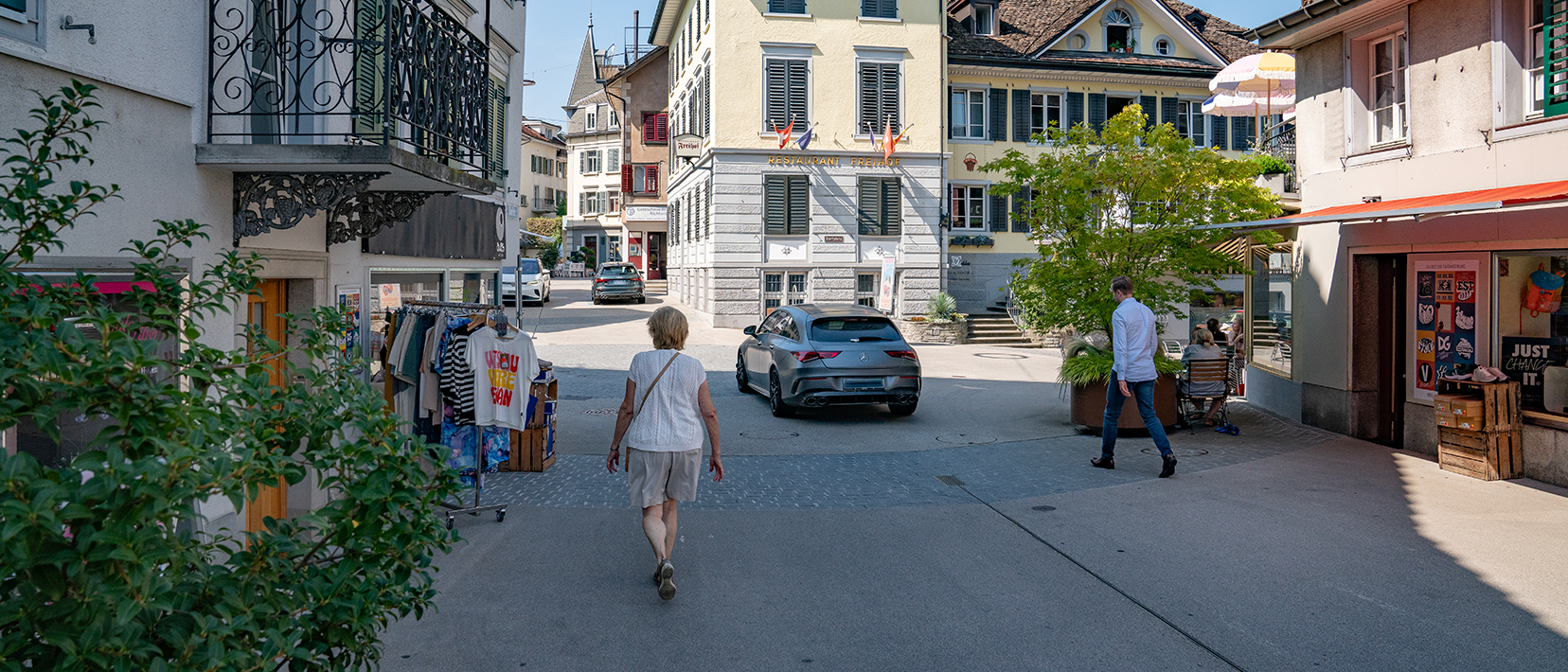 Fussgängerinnen und Fussgänger gehen durch einen Ortskern, in den Erdgeschossen befinden sich Geschäfte, im Hintergrund fährt ein Auto