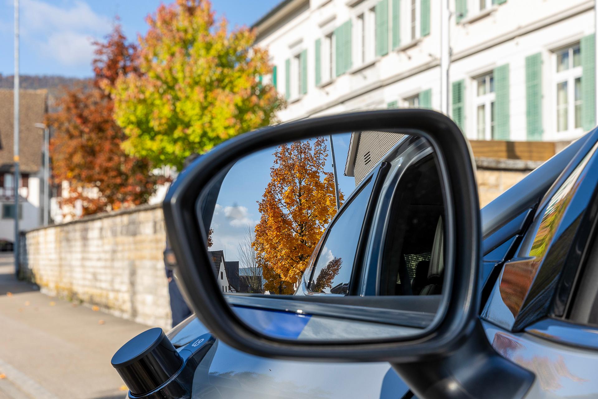Blick auf den linken Rückspiegel des selbstfahrenden Autos. Darin sieht man die Umgebung (u.a. einem herbstlichen Baum). 