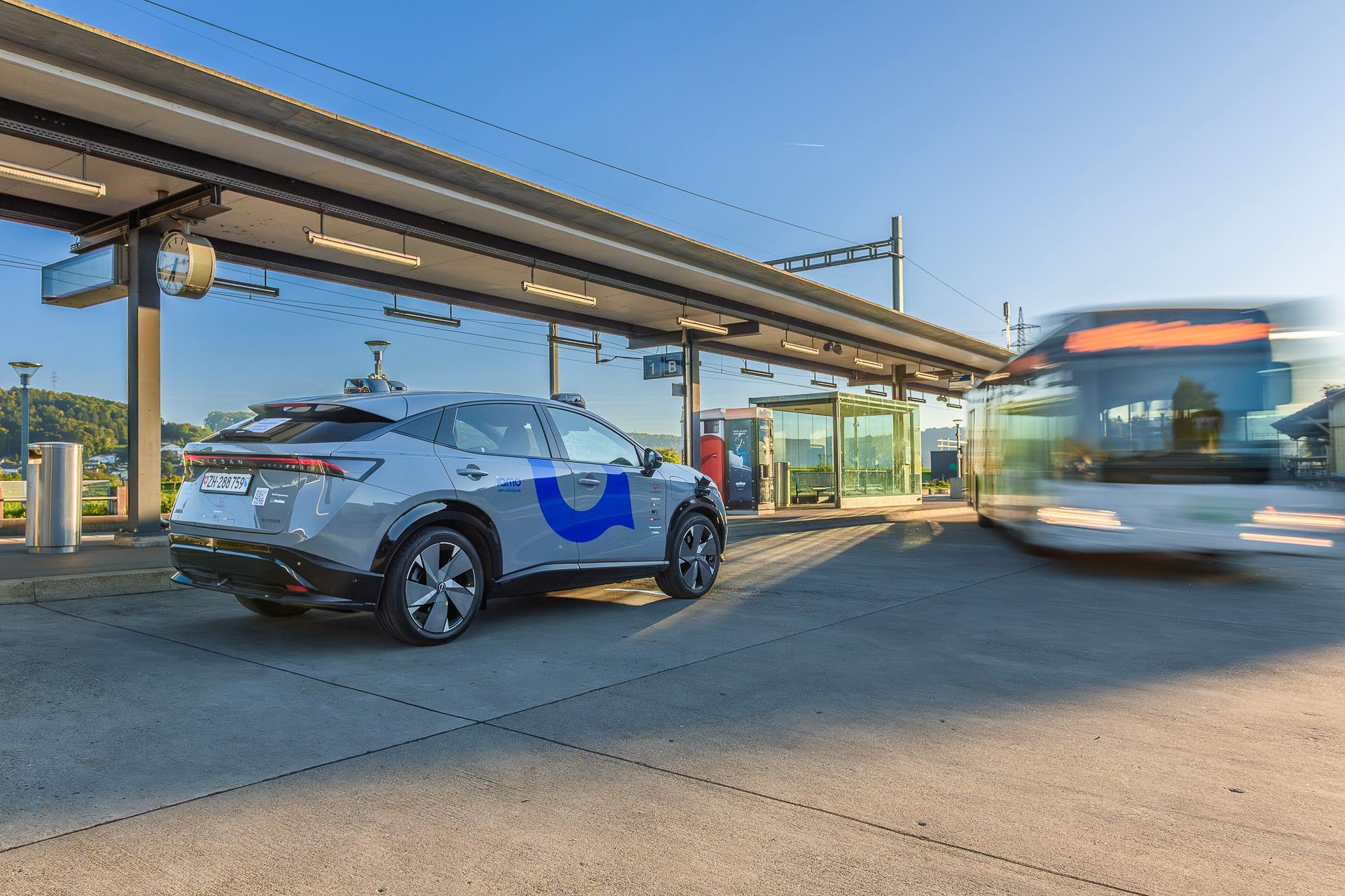 Ein selbstfahrendes steht am Bahnhof von Otelfingen. Im Hintergrund sind die Bahngleise und ein abfahrender Bus zu erkennen. 