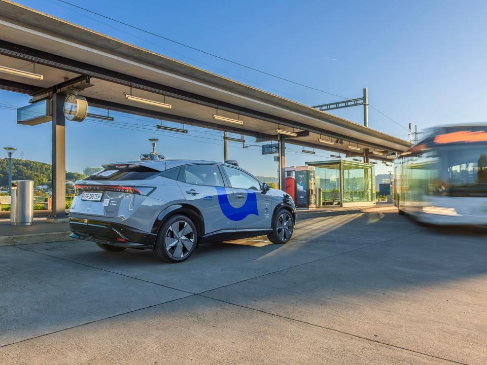 Ein selbstfahrendes steht am Bahnhof von Otelfingen. Im Hintergrund sind die Bahngleise und ein abfahrender Bus zu erkennen. 
