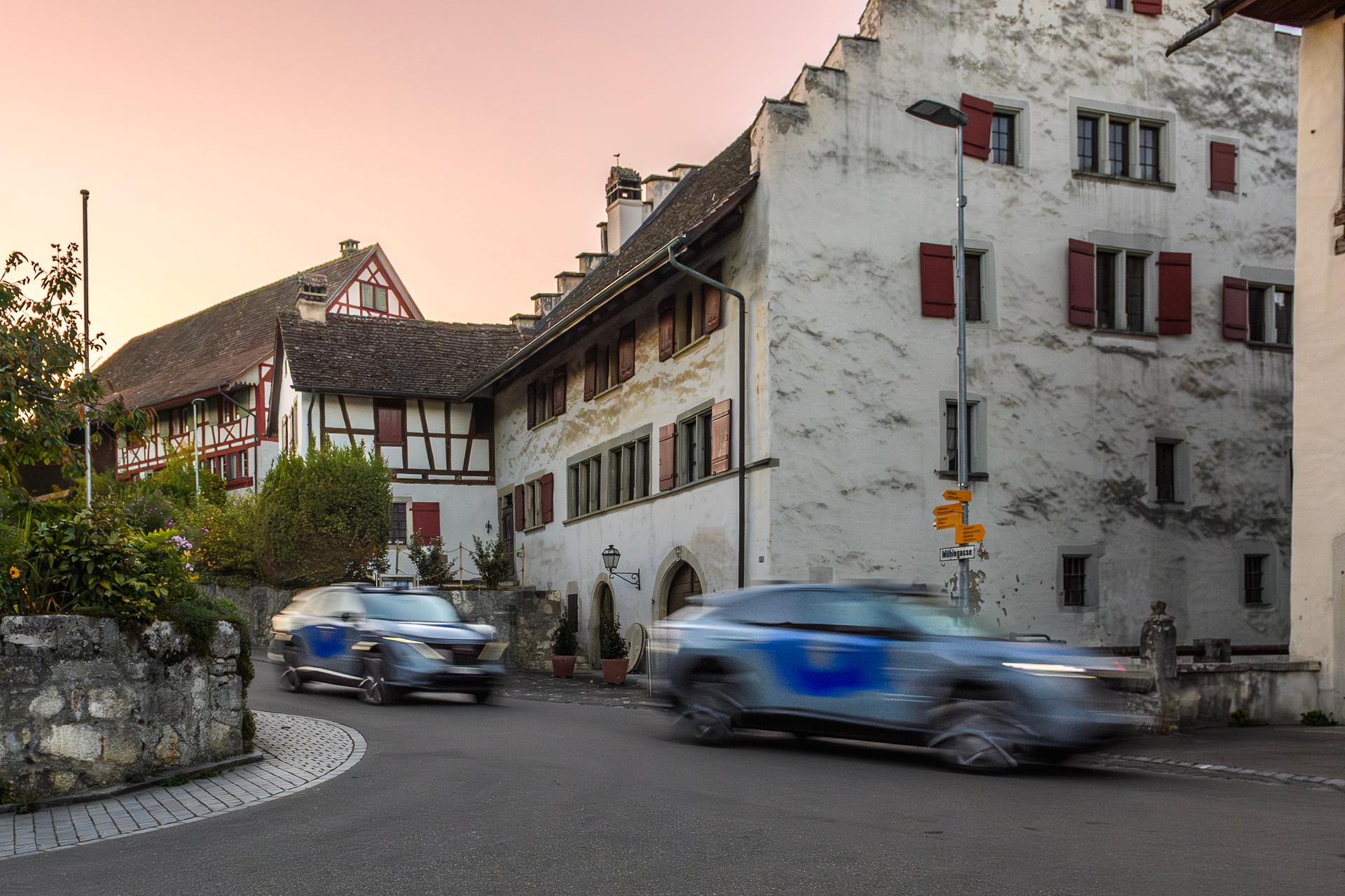 Zwei selbstfahrende Fahrzeuge fahren durch die Mühlegasse in Otelfingen. Im Hintergrund ist die historische Mühle zu sehen. 