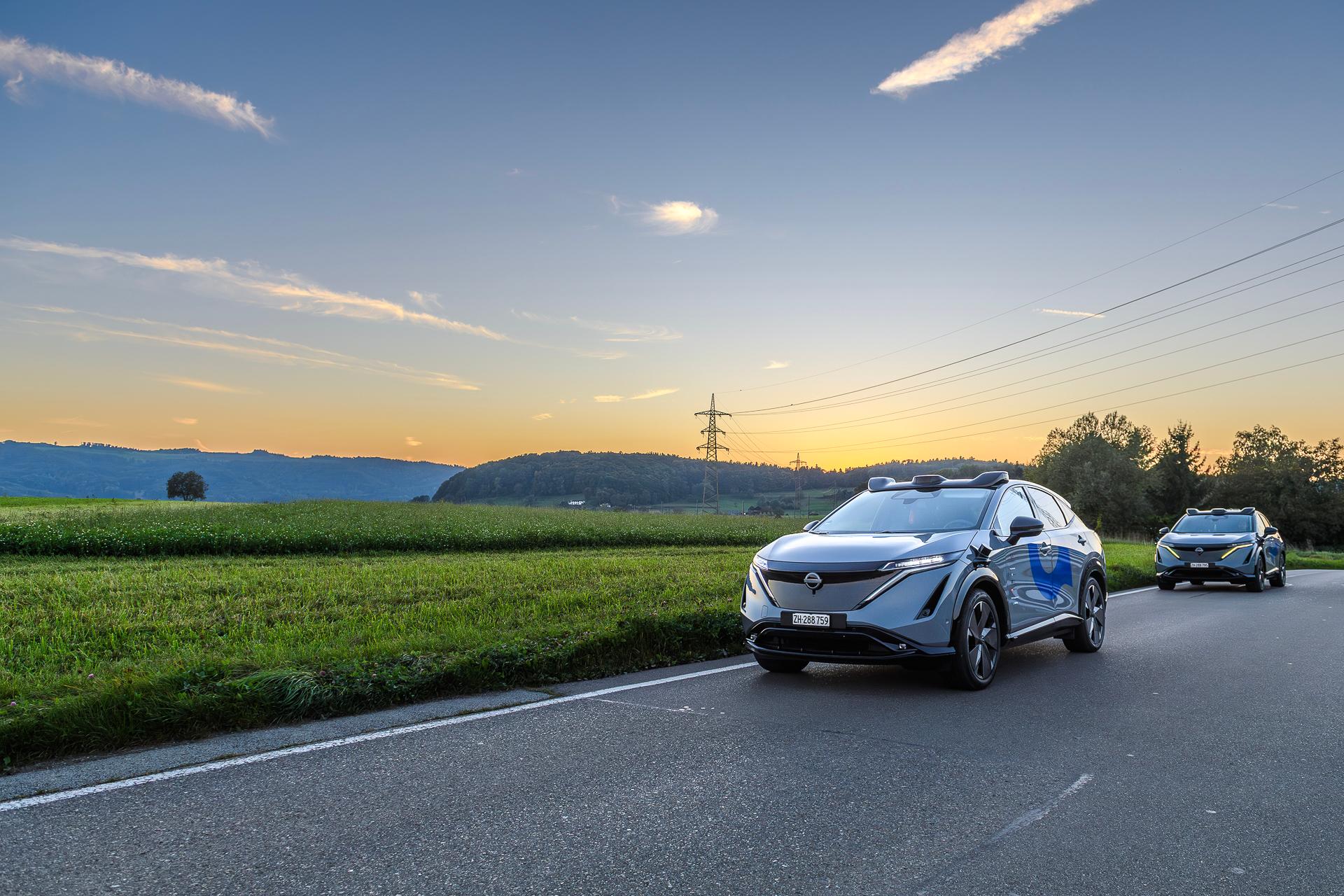 Zwei selbstfahrende Fahrzeuge fahren über eine Landstrasse im Zürcher Furttal. Der Himmel leuchtet bunt im Schein des Sonnenuntergangs.