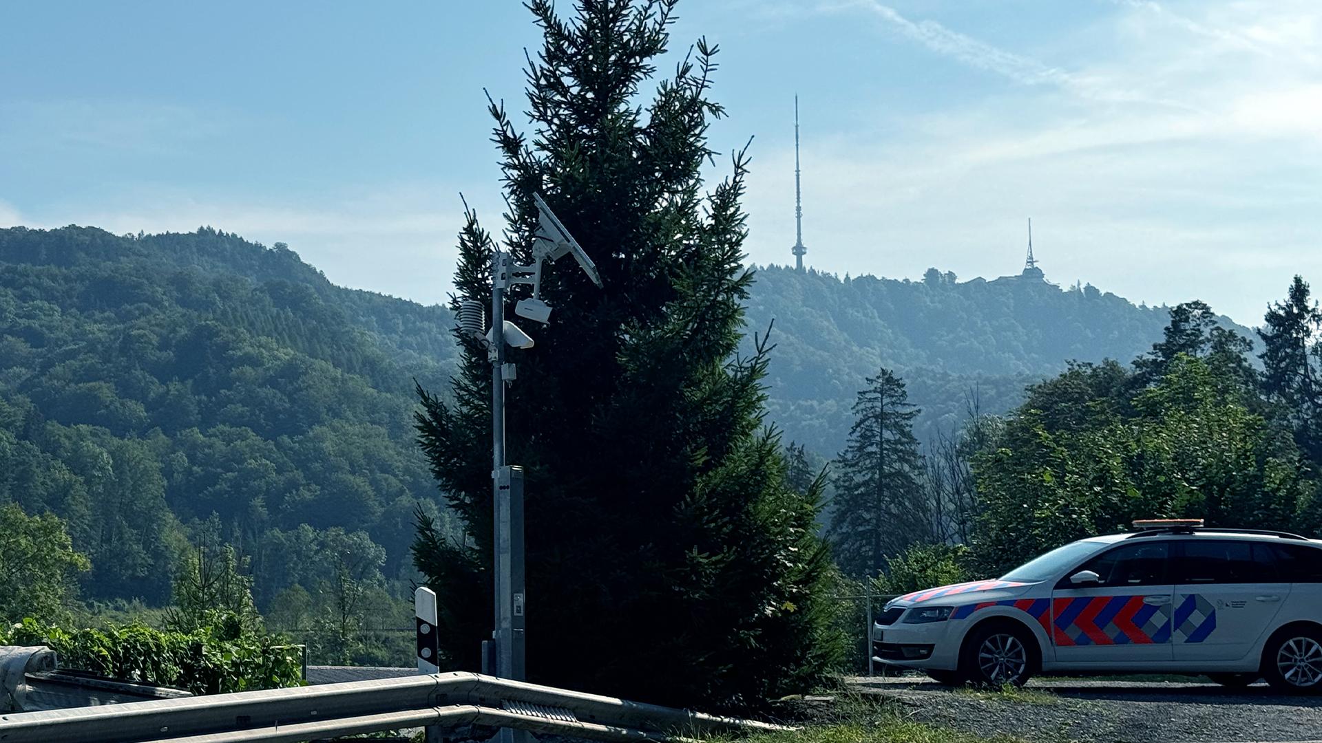 Frühwarnanlage neben einem Nadelbaum und Auto mit bergiger Landschaft im Hintergrund.