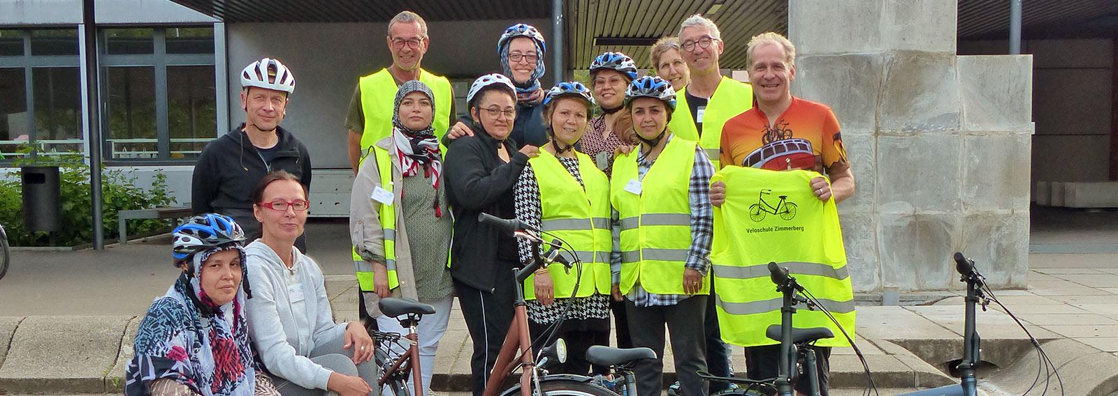 Gruppe Frauen und Männer, teils mit Velohelmen und Warnwesten, lächelt nach erfolgreichem Velokurs in die Kamera. Ein Mann hält eine Warnweste mit der Aufschrift «Veloschule Zimmerberg».