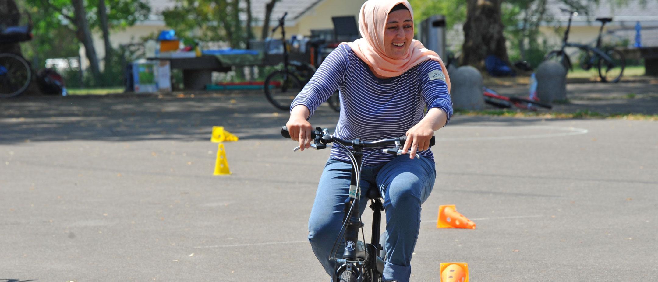 Lachende Frau mit Kopftuch und gestreiftem Oberteil fährt Fahrrad auf asphaltiertem Platz mit Pylonen.