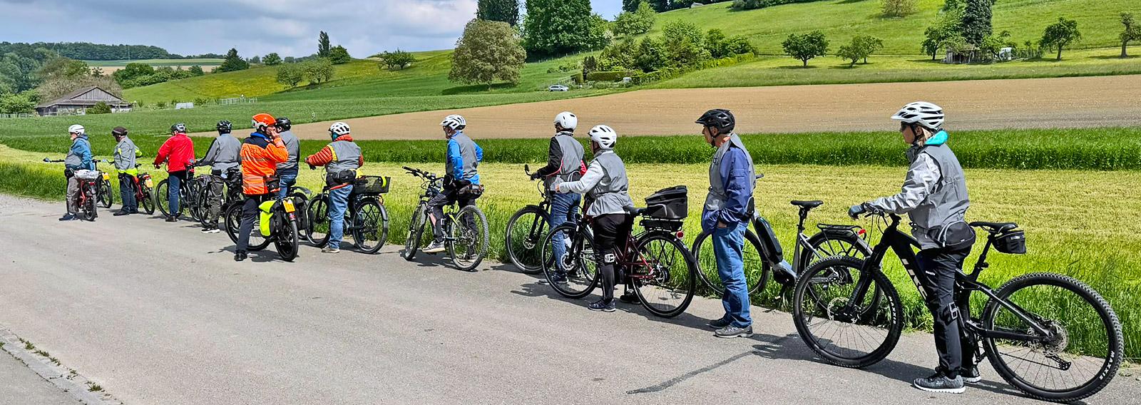 Gruppe von E-Bike-Fahrenden am Strassenrand wird vom Instruktor über die kommende Übung informiert. Im Hintergrund öffnet sich der Blick auf die grüne Hügellandschaft bei Bülach.