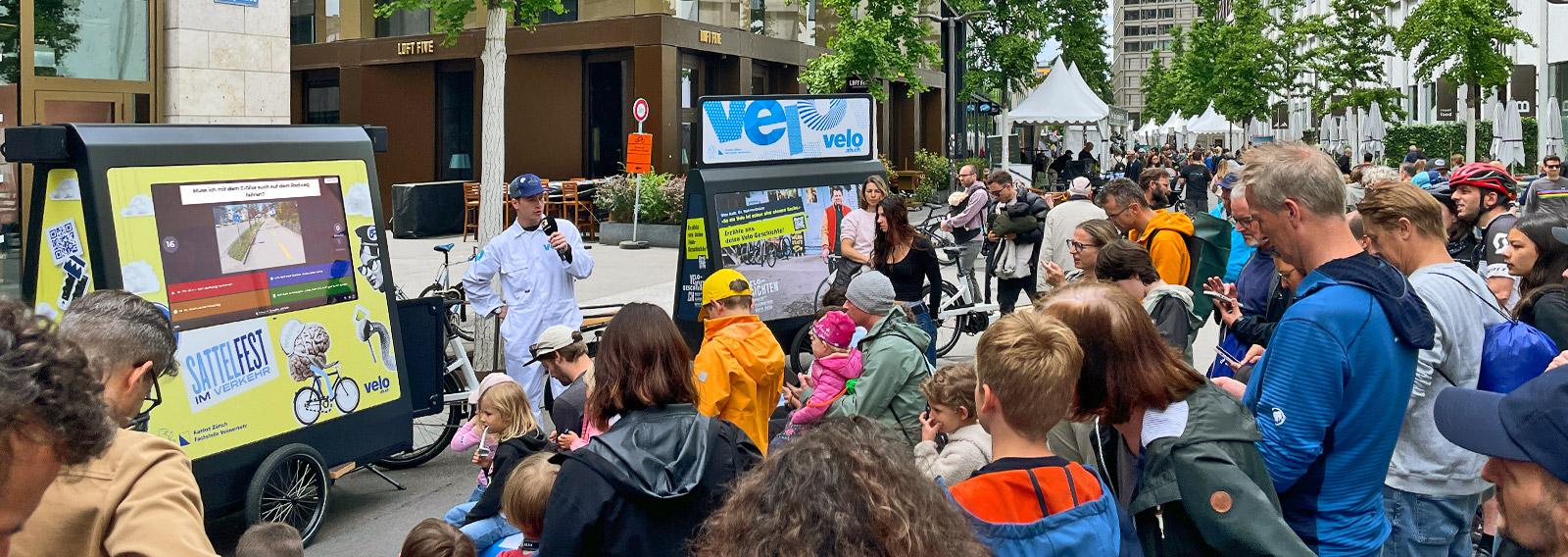 Ein Host in weissem Overall spricht am Stand der Fachstelle Veloverkehr an der Europaallee zum Publikum, das sich vor einem grossen Bildschirm versammelt hat.
