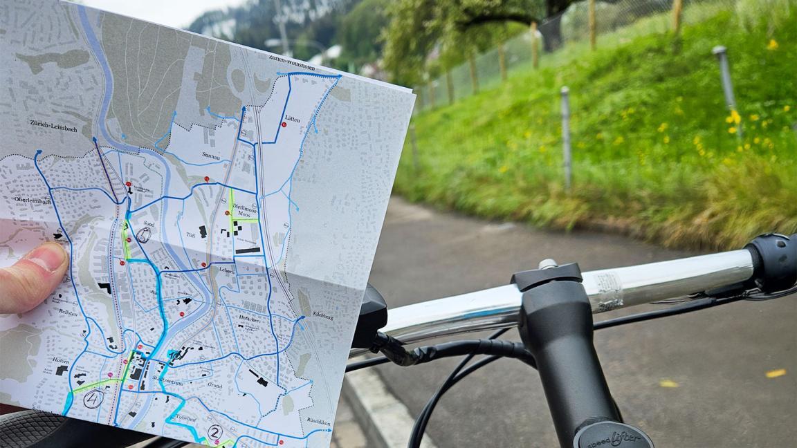 Jemand hält eine Kopie des Velonetzplans der Gemeinde Adliswil in der Hand. Darauf sind gelb und blau markierte Routen zu sehen. Im Hintergrund des Plans sind ein Velolenker und eine asphaltierte Strasse zu sehen.