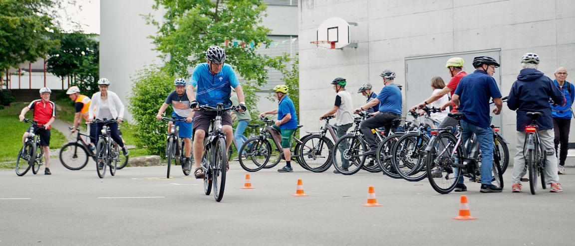 Ein Slalomkurs ist am Boden eines Schulhausplatzes markiert und ide Teilnehmenden stehen mit ihren E-Bikes bereit. Eine Person fährt am Parcour vorbei