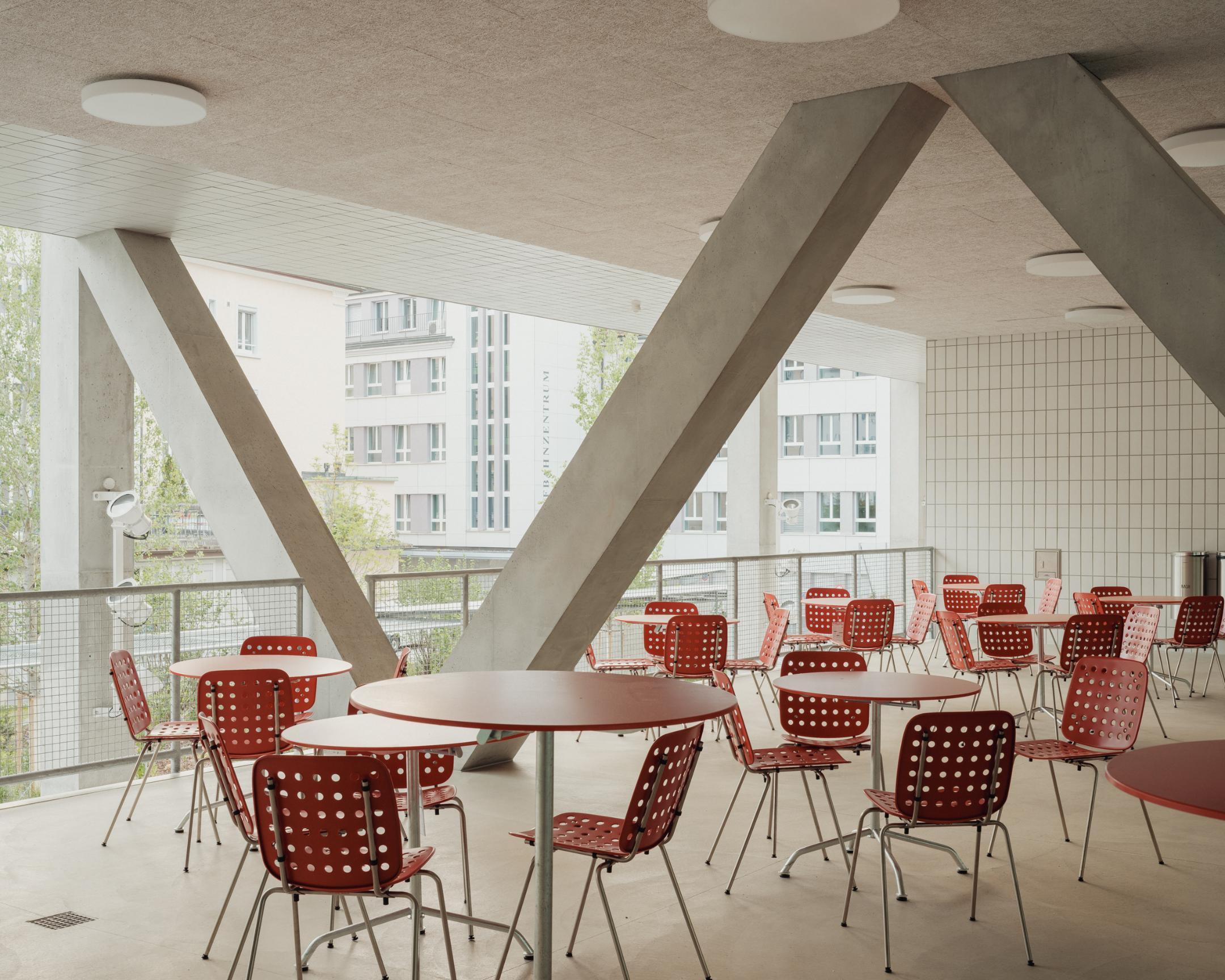 Überdachte Terrasse mit roten Stühlen und runden Tischen, gestützt von diagonalen Betonträgern.