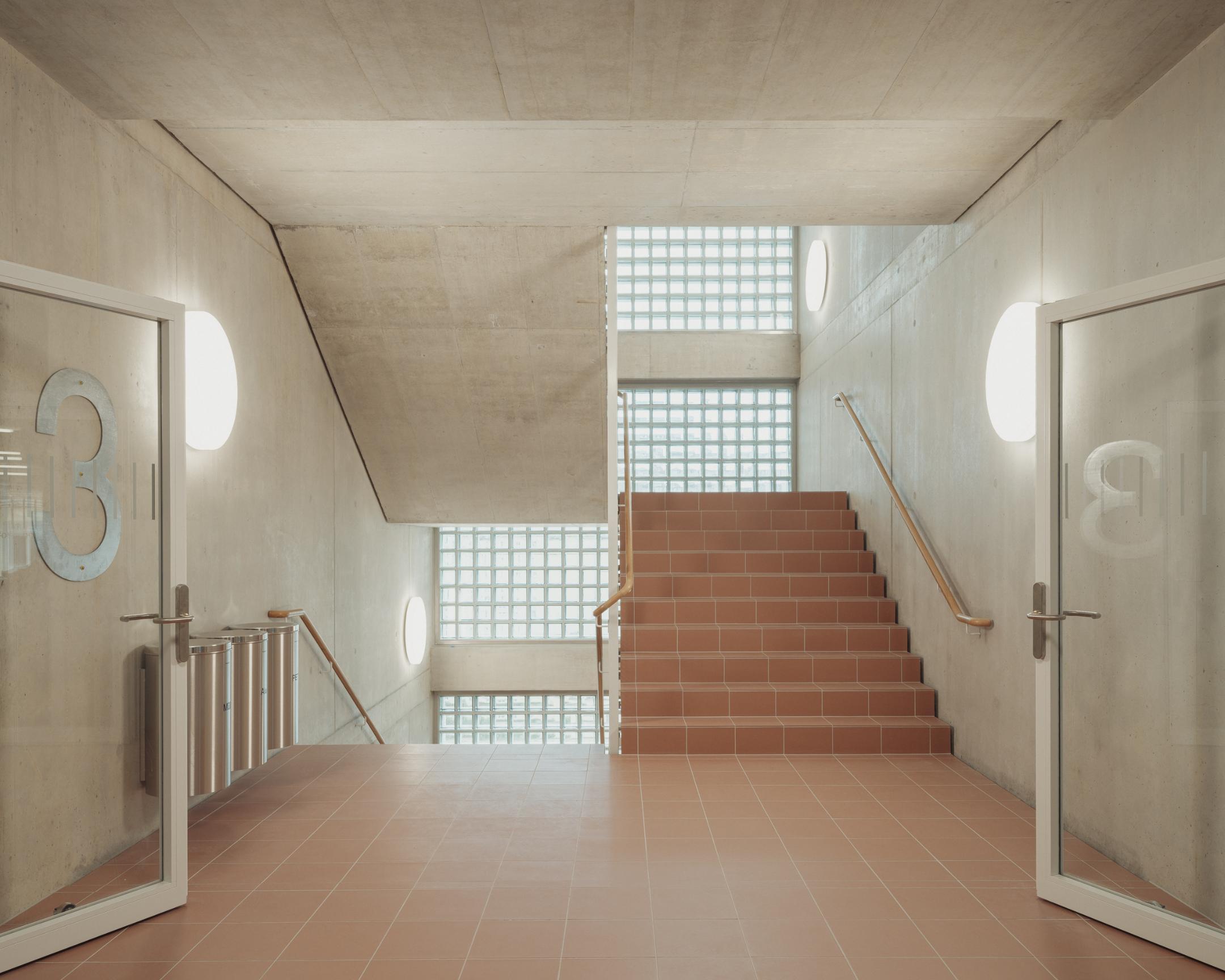Innenansicht eines Treppenhauses mit Betonwänden, rötlichen Bodenfliesen und Glasbausteinen als Lichtquelle.