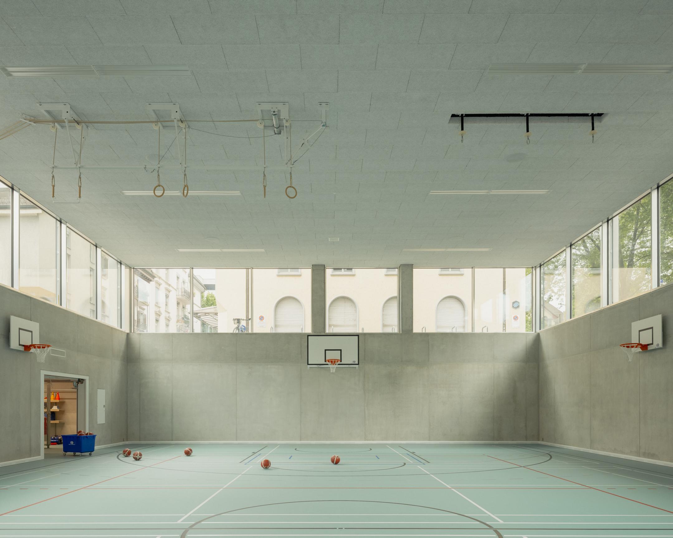 Innenansicht einer modernen Turnhalle mit Basketballkörben an den Wänden, mehreren Bällen auf dem Spielfeldboden und hohen Fenstern, die Tageslicht hereinlassen.