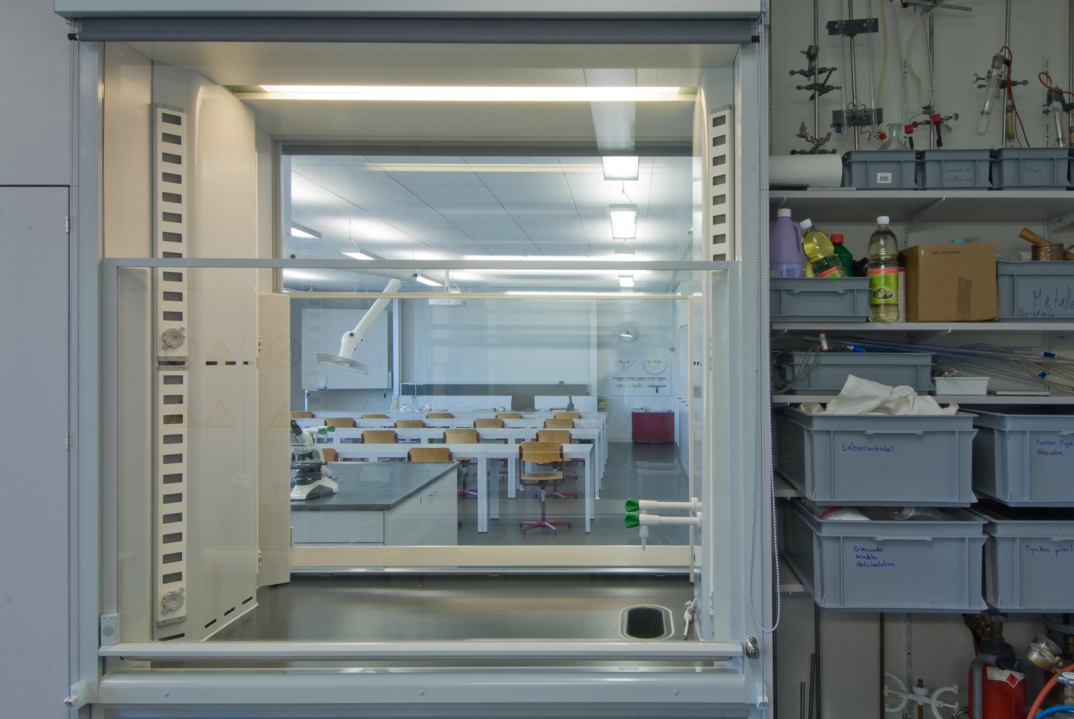 Ansicht aus einem Raum mit Kunststoffkisten, welche mit diversem Material wie Papier, Kunststoffflaschen und ähnlichem befüllt sind. Durch ein Innenfenster sieht man in einen Klassenraum.