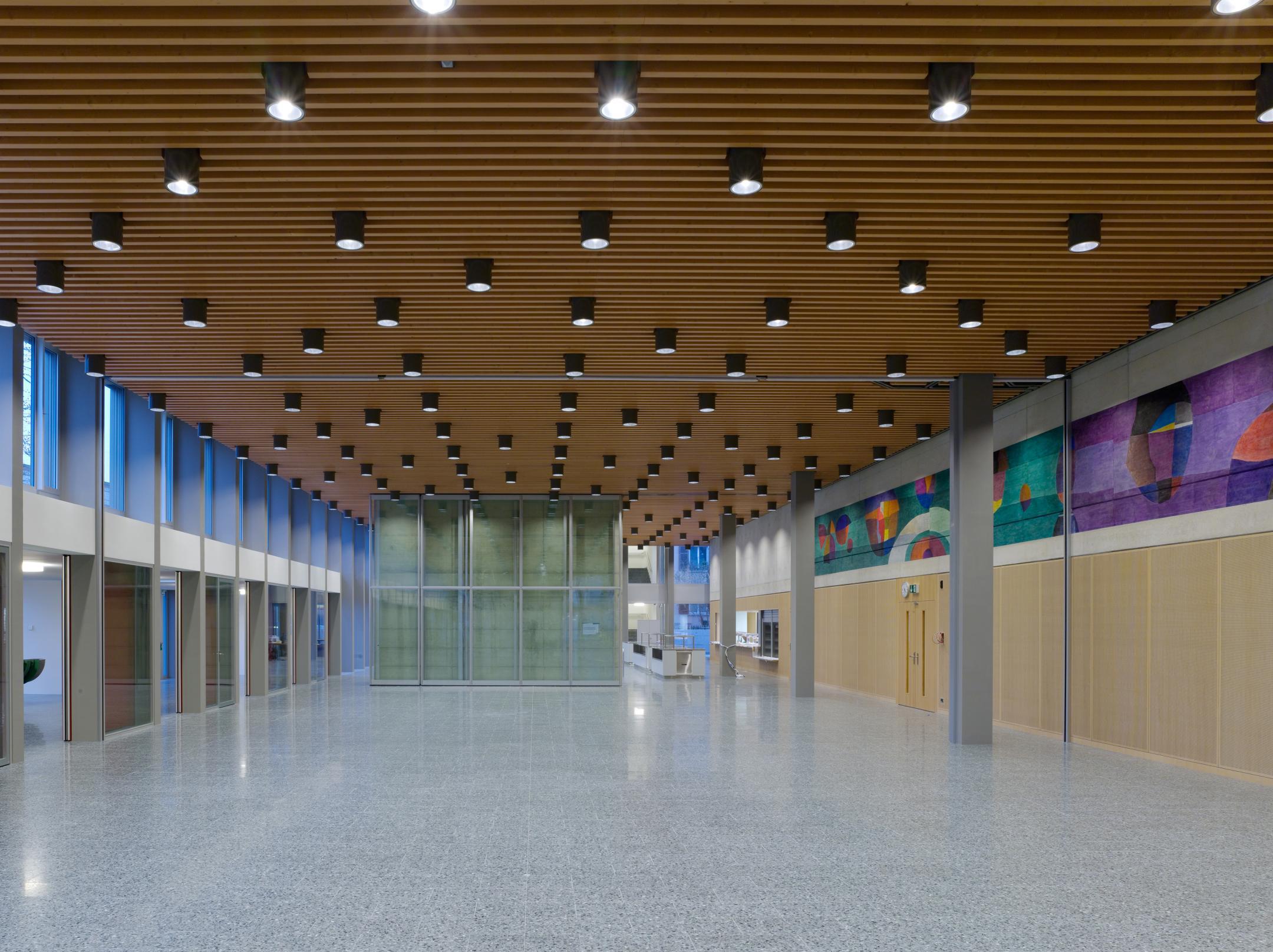 Innanansicht eines grossen Raumens, auf mehreren Seiten offen ist. Auf der rechten, geschlossenen Seite ist über der mannhohen Holzverkleidung ein farbiges gemaltes Band aus geometrischen Figuren zu sehen.