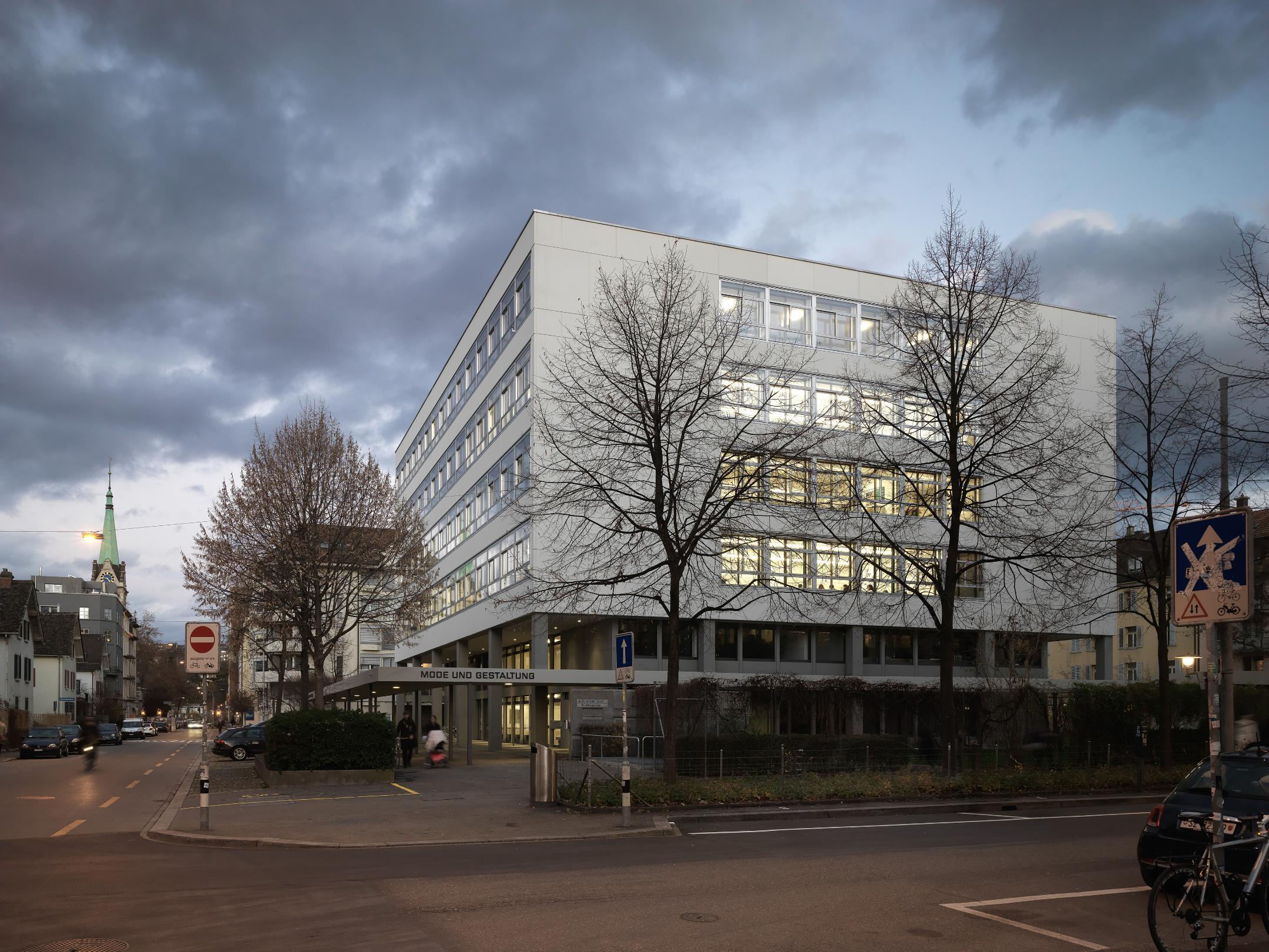 Kubisches Gebäude in der Abenddämmerung an Strassenkreuzung. Die Fenster sind beleuchtet.