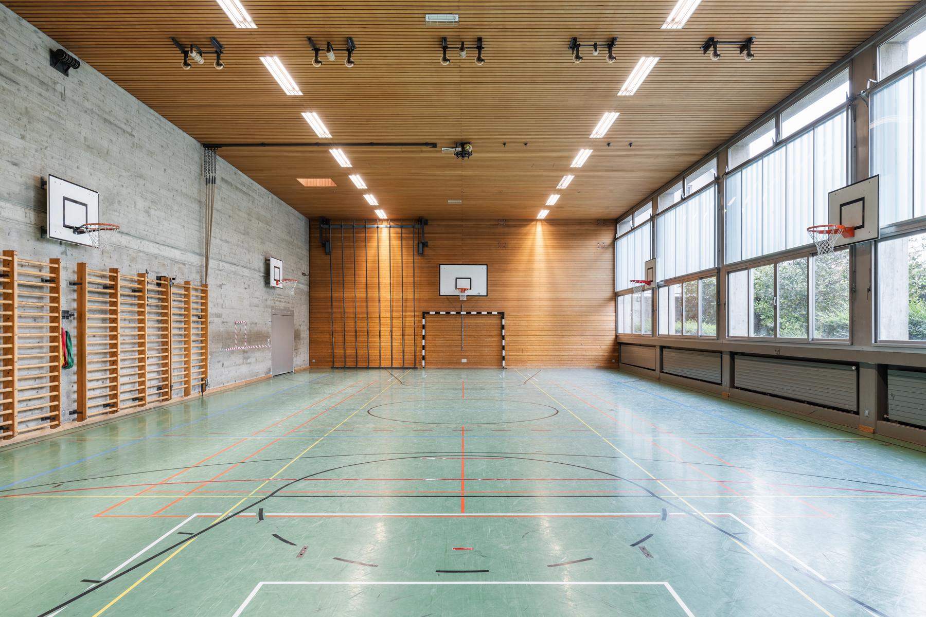 Symmetrische Innenperspektive der bestehenden Sporthalle welche viel Holzverkleidungen und Beton aufzeigt.