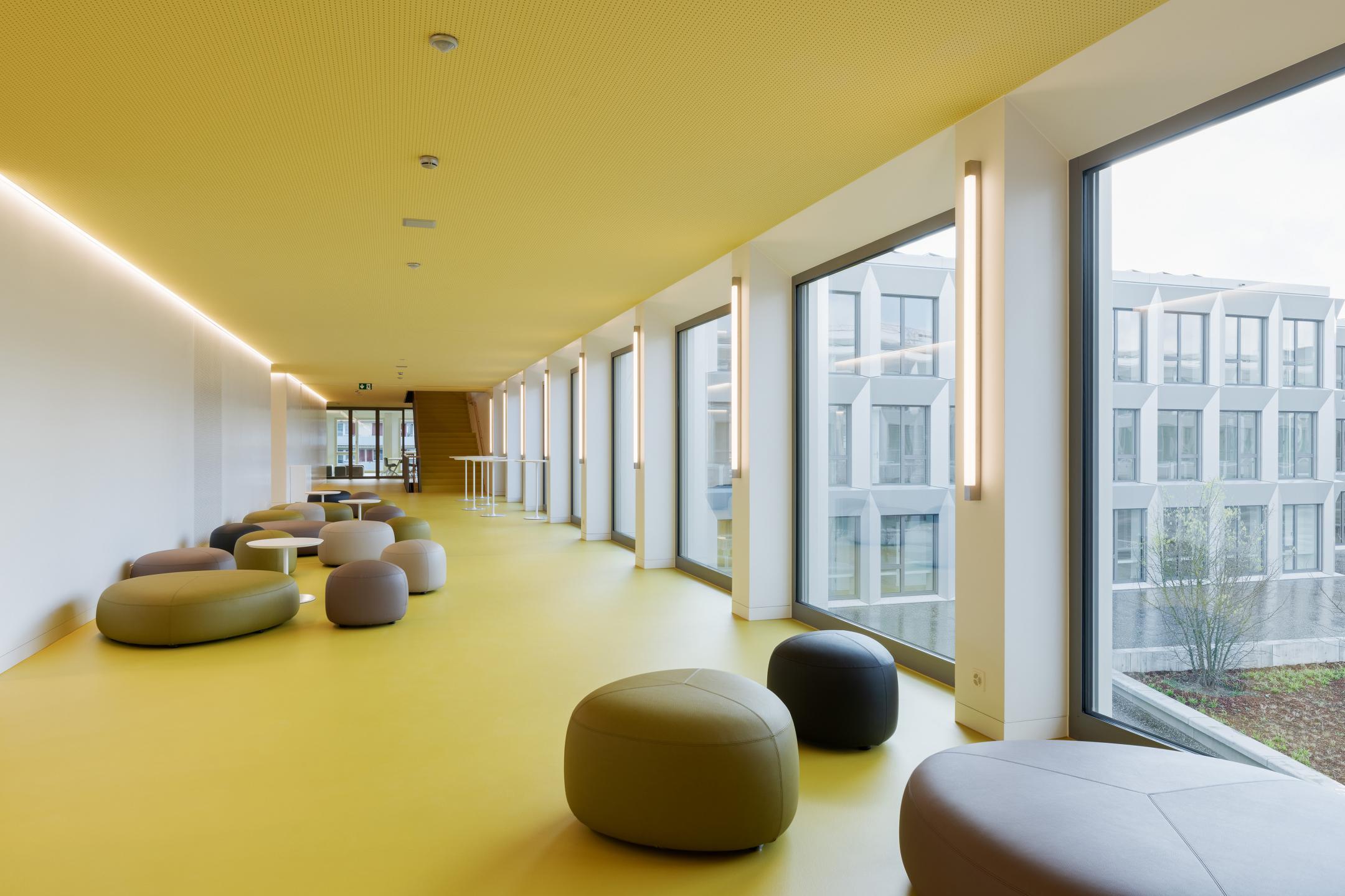 Innenansicht eines breiten Korridors. Die Wände sind in hellem weiss, Boden und Decke in Gelb gehalten. Um kleine Tische sind Sitzgelegenheiten in Form von runden Sitzsäcken gruppiert.