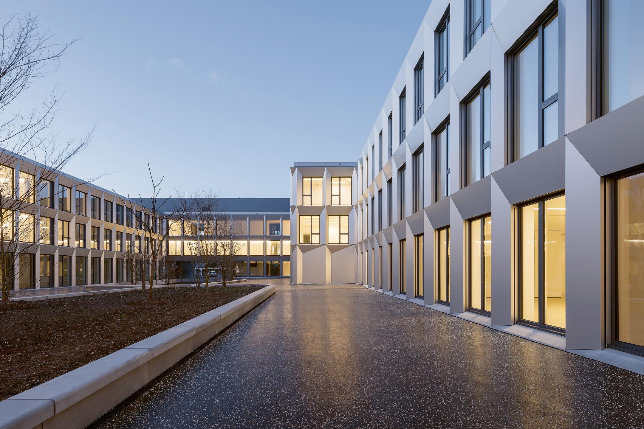 Innenhof in der Abenddämmerung. Das Gebäude ist Beleuchtet und umfasst den Innenhof komplett. In der Mitte des Innenhofes ist eine humusierte Fläche zu sehen. Sie ist mit jungen Bäumen bepflanzt.