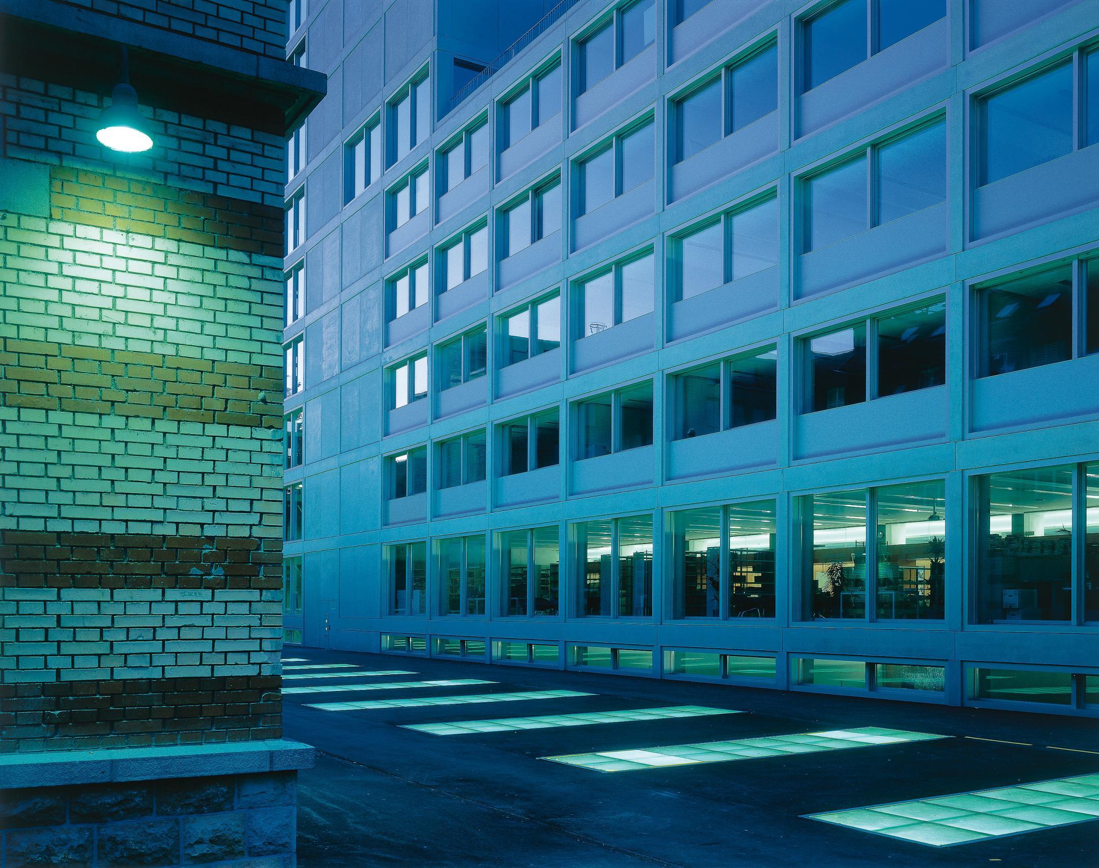 Ansicht in der Abenddämmerung an ein Gebäude mit grossen Glasflächen. Der Platz vor dem Gebäude ist durch in den Boden eingelassene Lichtbänder unterteilt.