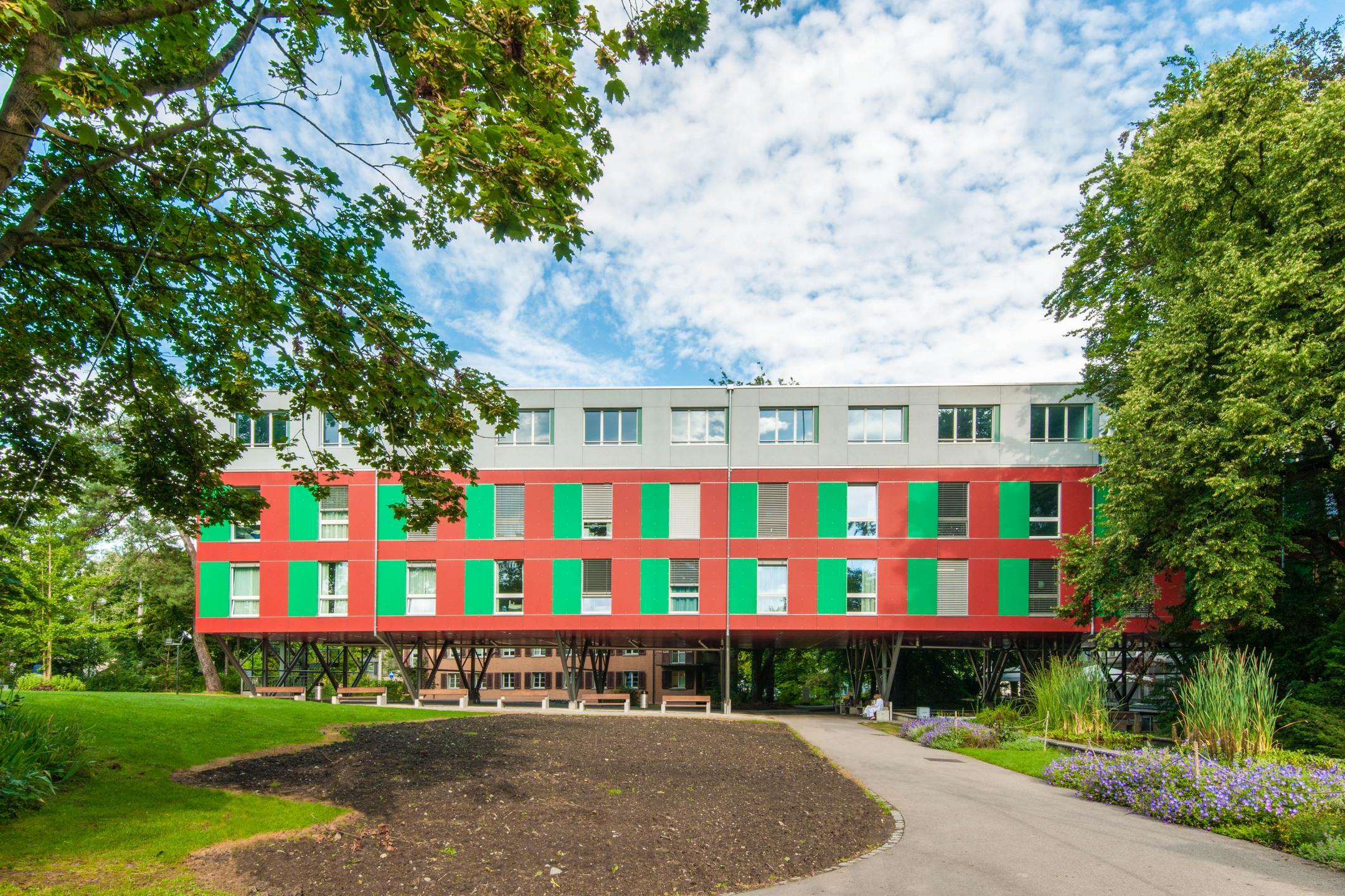 Gebäudekomplex in Gartenanlage. Das dreigeschossige Gebäude ist mit Flachdachversehen. Die untern zwei Geschosse sind in rot, das oberste Geschoss in weiss gehalten.