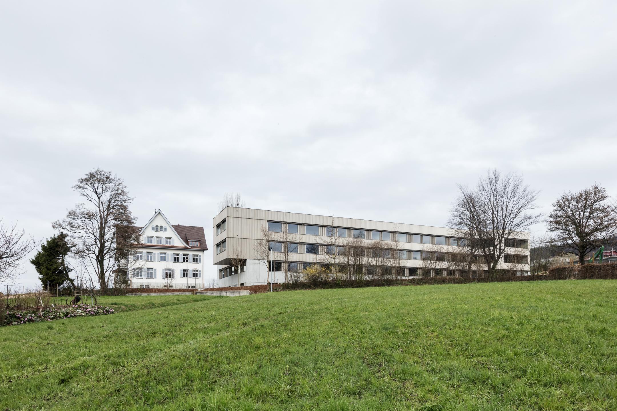 Zwei Gebäude auf einer Wiese. Links ein älteres Gebäude mit Steildach, links ein neues Gebäude mit Flachdach.