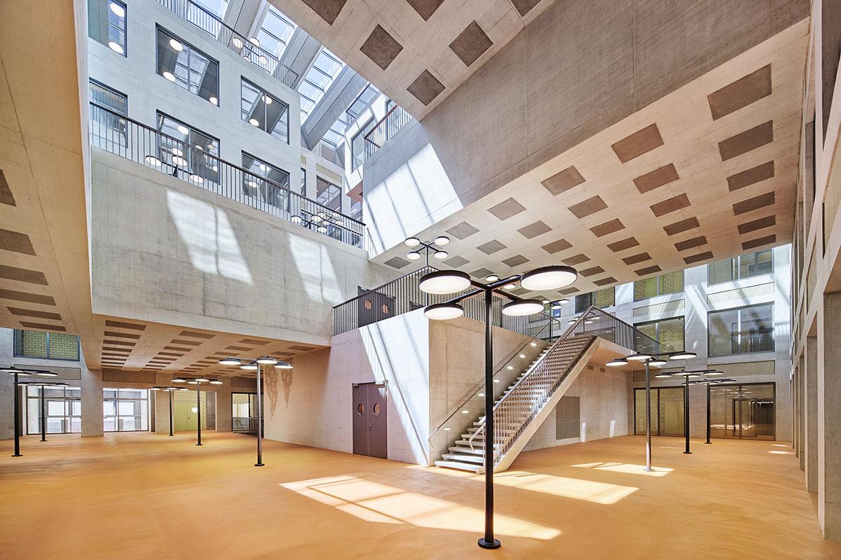 Mit den Strassenlaternen sowie dem reduzierten Farb- und Materialkonzept bringt das lichtdurchflutete Atrium den Aussenraum in das Gebäude. Die in die Betondecken eingelassenen Akustikpaneele übernehmen analog das Bild der Lochfassaden im Innenhof. Die im Innenhof reinkragenden Unterrichtsräume sind spielerisch versetzt und aufeinandergestapelt. Diese Volumen lassen den inneren Bereich zu einer begehbaren Raumskulptur werden. Sie erstreckt sich vom ersten Untergeschoss bis unter das alles überspannende Glasdach.