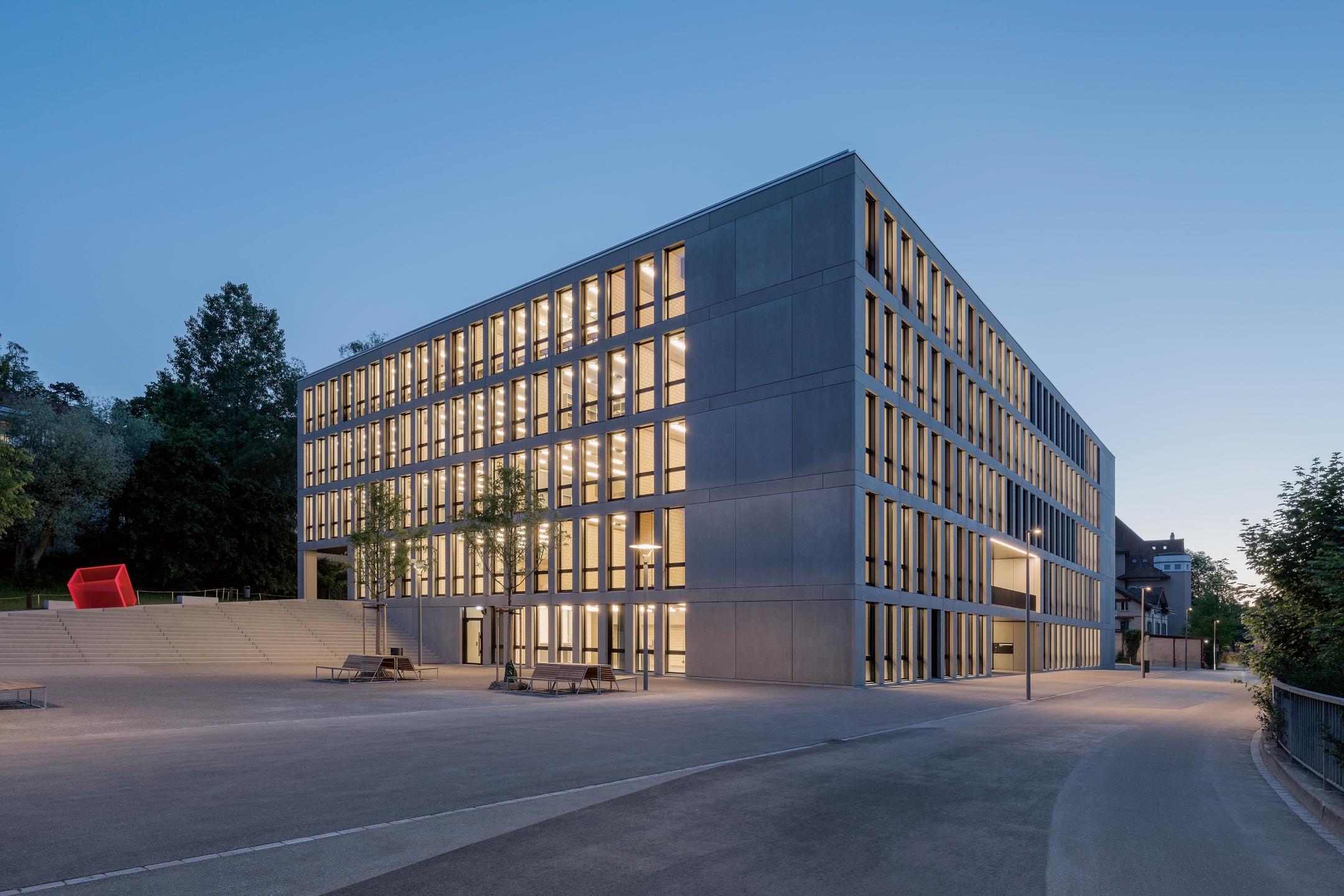 Kubischer Neubau bei Abenddämmerung mit beleuchteten Fenstern. Treppenaufgang auf einer Gebäudeseite und darüber ein magentafarbener, transparenter Kubus.