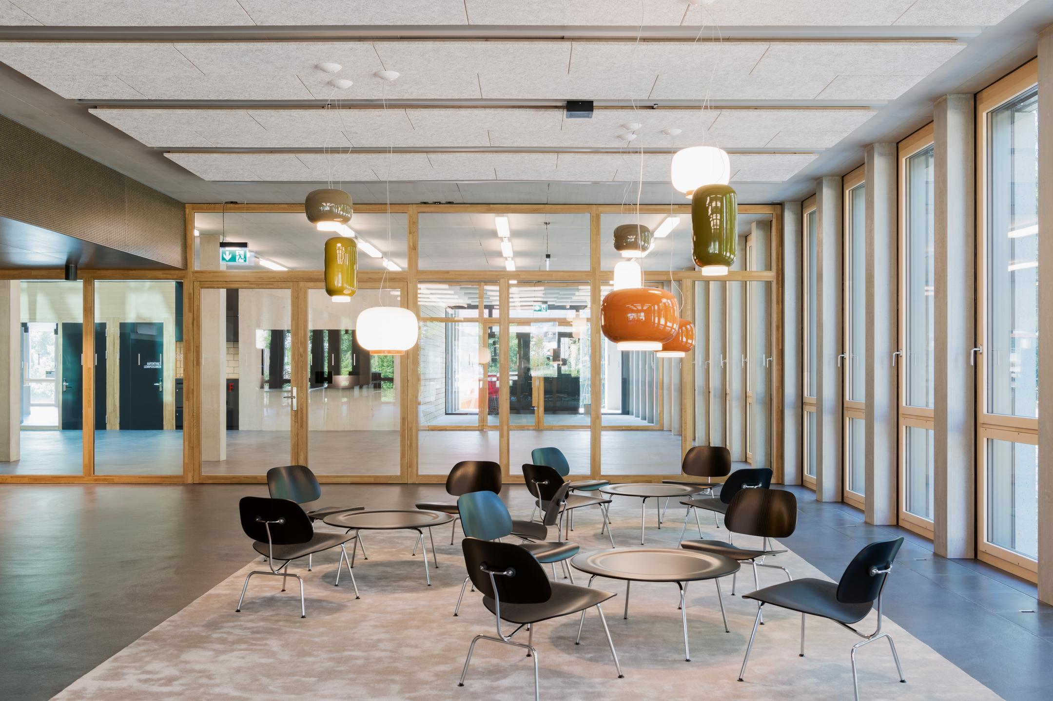 Sitzgelegenheit mit kleinen Tischen und Stühlen im hellen Korridor. Die Wände sind alle verglast. Von der Decke hängen farbige Leuchter.