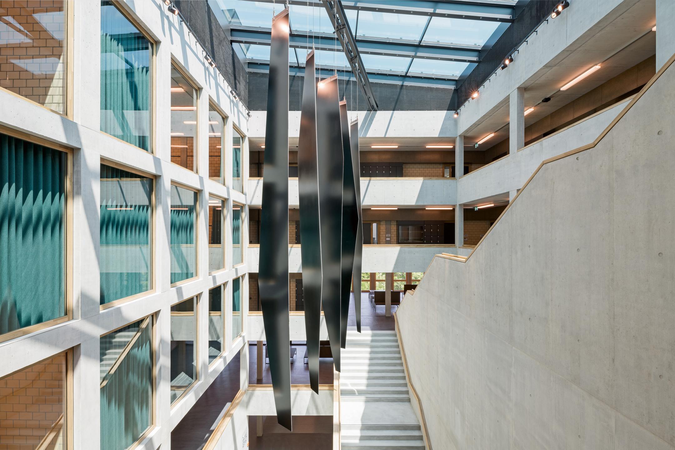 Überdachter Innenhof mit Glasfenstern, offenem Treppenaufgang und Laubengang. Metallstreifen hängen von Glasdach in der Mitte des Hofes.