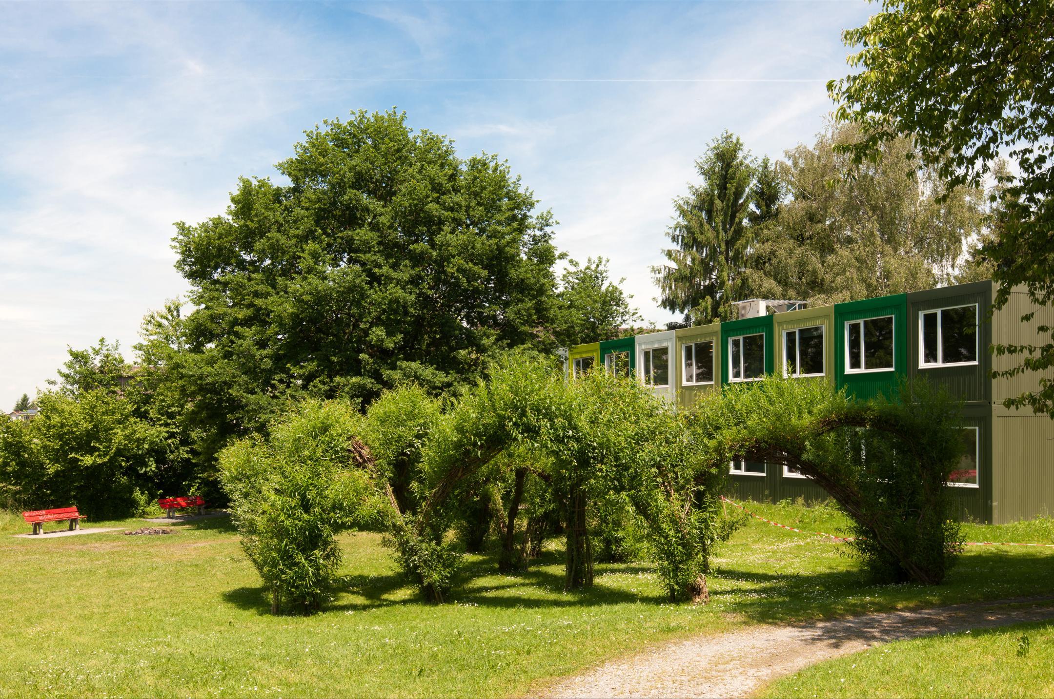 Parklandschaft im Sommer mit Bäumen, Büschen und roten Sitzbänken. Im Hintergrund steht ein in verschiedenen grüntönen gehaltenes, einfaches Haus.