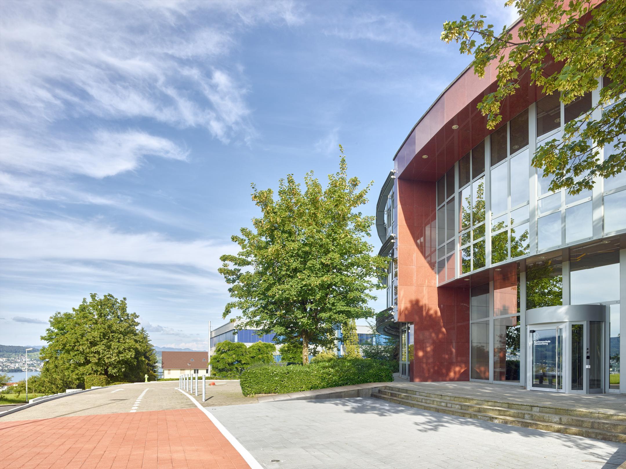 Das Bild zeigt die bestehende, dreigeschossige Nordfassade der Mietliegenschaft an der Steinacherstrasse, die für die neue Kantonsschule Zimmerberg instandgesetzt wurde. Die architektonische Sprache im Haupteingangsbereich orientiert sich mit der gerundeten Fassade, den rötlichen Kunststeinplatten und den raumhohen Verglasungen an der Postmoderne.