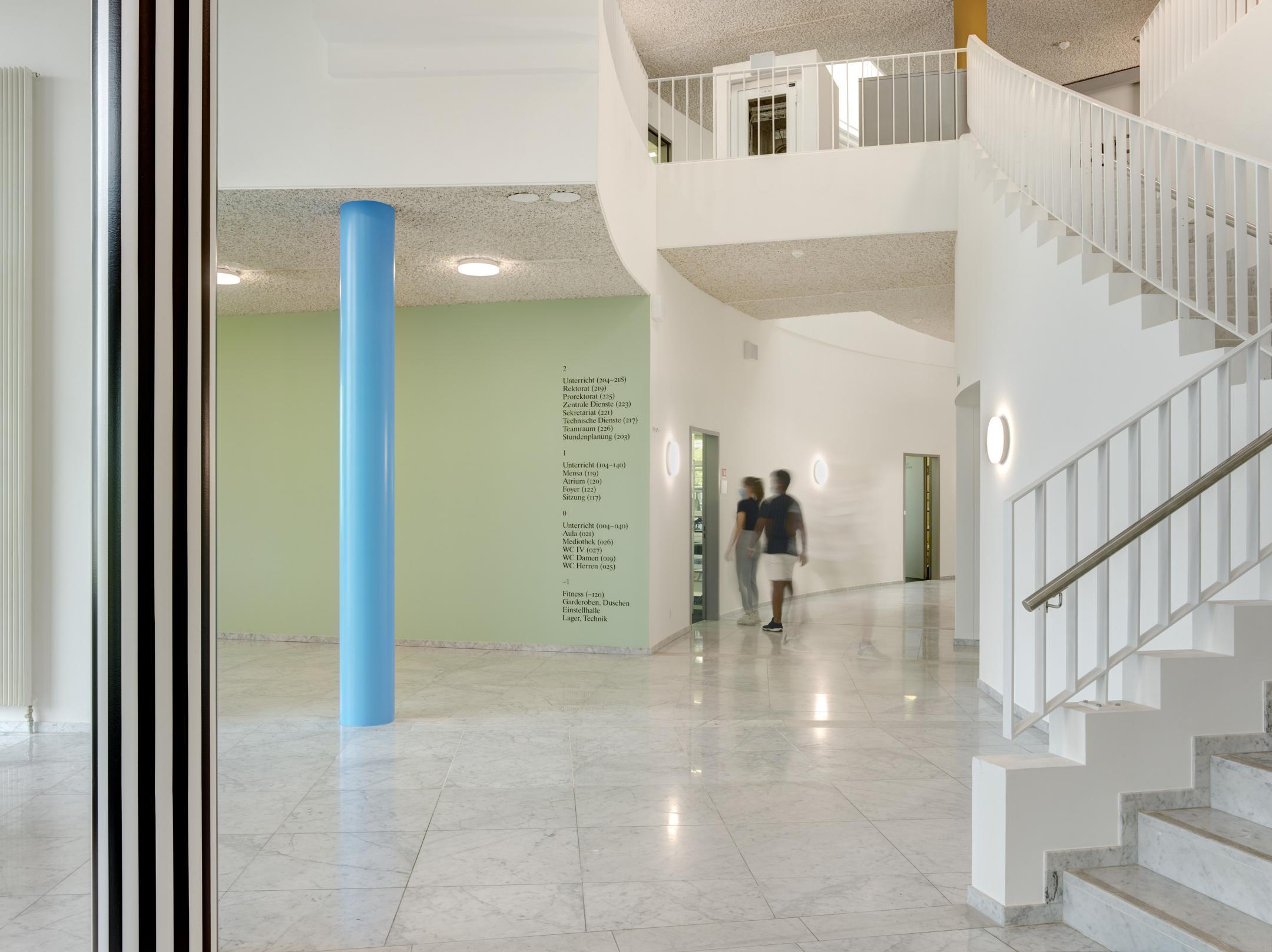 Das Bild zeigt den Eingangsbereich mit seiner repräsentativen Erschliessungshalle und Doppeltreppe. Die räumlichen Qualitäten werden mittels des Signaletik- und Farbkonzeptes gestärkt. Angelehnt an die Designideen der Gruppe Memphis werden die Säulen und gewisse Wandflächen mit Farben, Beschriftung und «Pattern» als kreativer, aber auch subtile Ergänzung zur bestehenden Gebäudesubstanz behandelt.