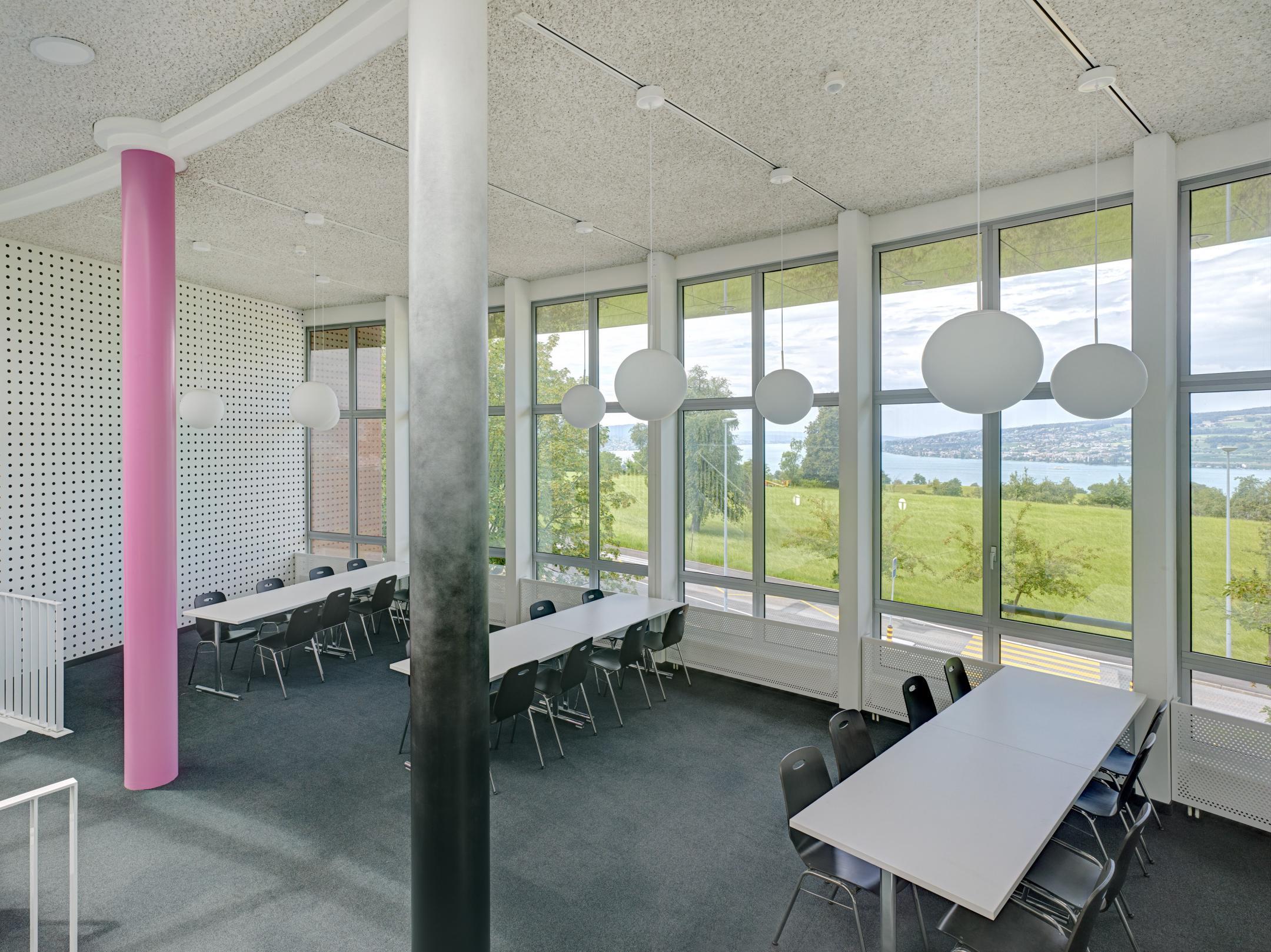 Das Bild zeigt den prägenden Aufenthaltsbereich im Zwischengeschoss mit inspirierenden Ausblicken auf den Zürichsee. Die Fläche wird als Begegnungsraum, aber auch als Ergänzungsbereich mit Gruppenarbeitstische belegt.