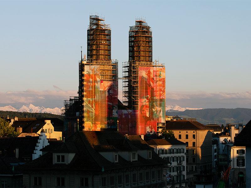 Fernaufnahme des Zürcher Grossmünsters im warmen Abendlicht, dessen eingerüstete Türme mit bunten, kunstvoll bedruckten Planen verkleidet sind. Im Hintergrund erstreckt sich die Silhouette der schneebedeckten Alpen unter einem hellen Himmel.