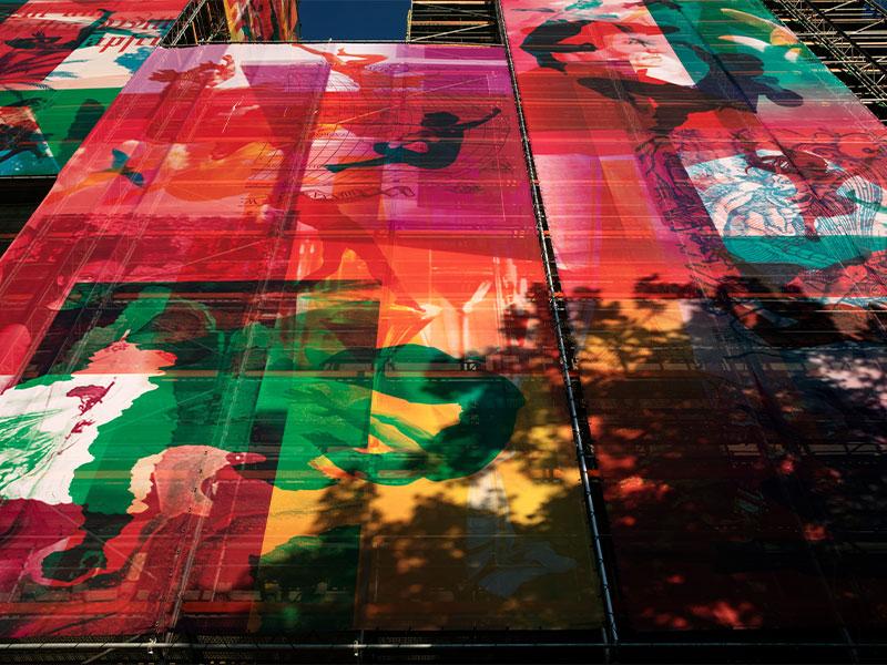 Detailansicht einer grossflächigen, halbtransparenten Kunst-Plane an einem Baugerüst. Zu sehen ist eine farbintensive Collage aus Rot-, Violett- und Grüntönen mit angedeuteten menschlichen Silhouetten und grafischen Mustern, auf die der Schatten von Bäumen fällt.