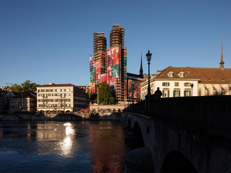 Blick über die Limmat in Zürich auf das eingerüstete Grossmünster mit seinen farbigen Kunst-Planen. Im Vordergrund ist die Münsterbrücke im Schatten zu sehen, während die Sonne auf der Wasseroberfläche reflektiert und die Altstadtgebäude beleuchtet.