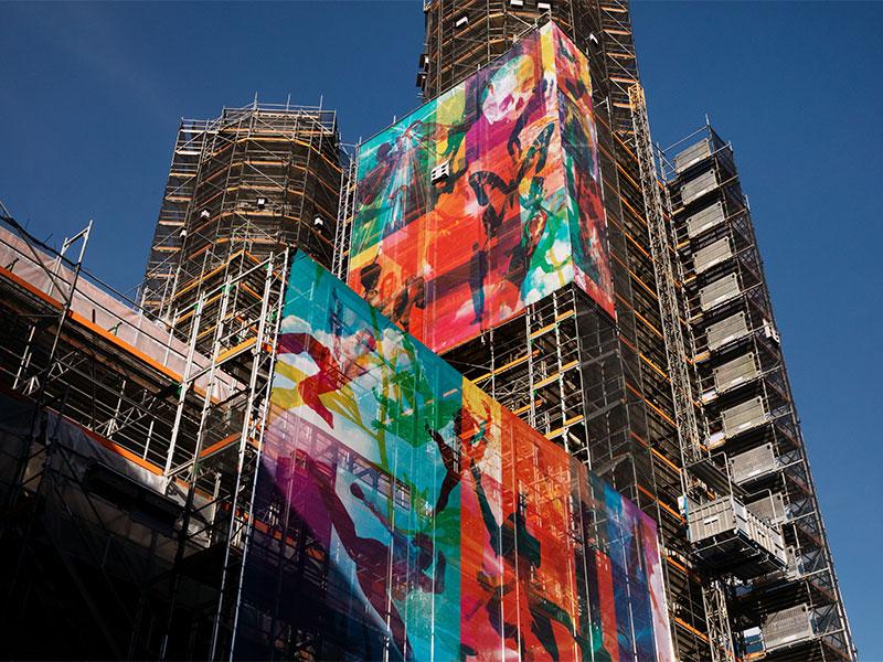 Nahaufnahme der beiden in Gerüste gehüllten Türme des Grossmünsters in Zürich unter blauem Himmel. Grosse, farbenfrohe Kunst-Planen mit abstrakten Figuren und lebendigen Farbverläufen in Orange, Rot, Grün und Blau verdecken Teile der Baustelle.