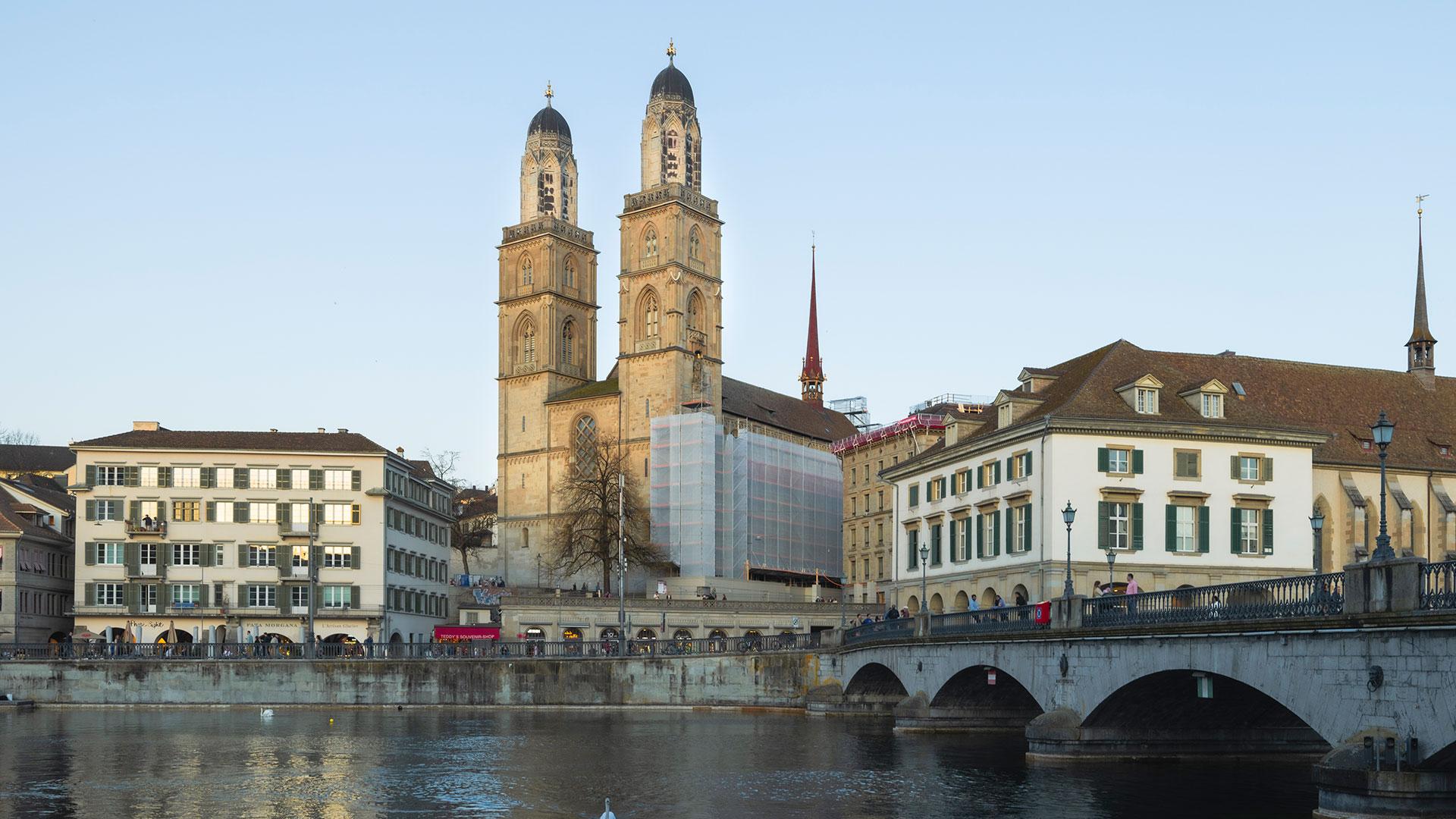 Das Grossmünster mit seinen markanten Doppeltürmen überragt die Limmat in Zürich, umgeben von historischen Gebäuden unter einem klaren, blauen Himmel.