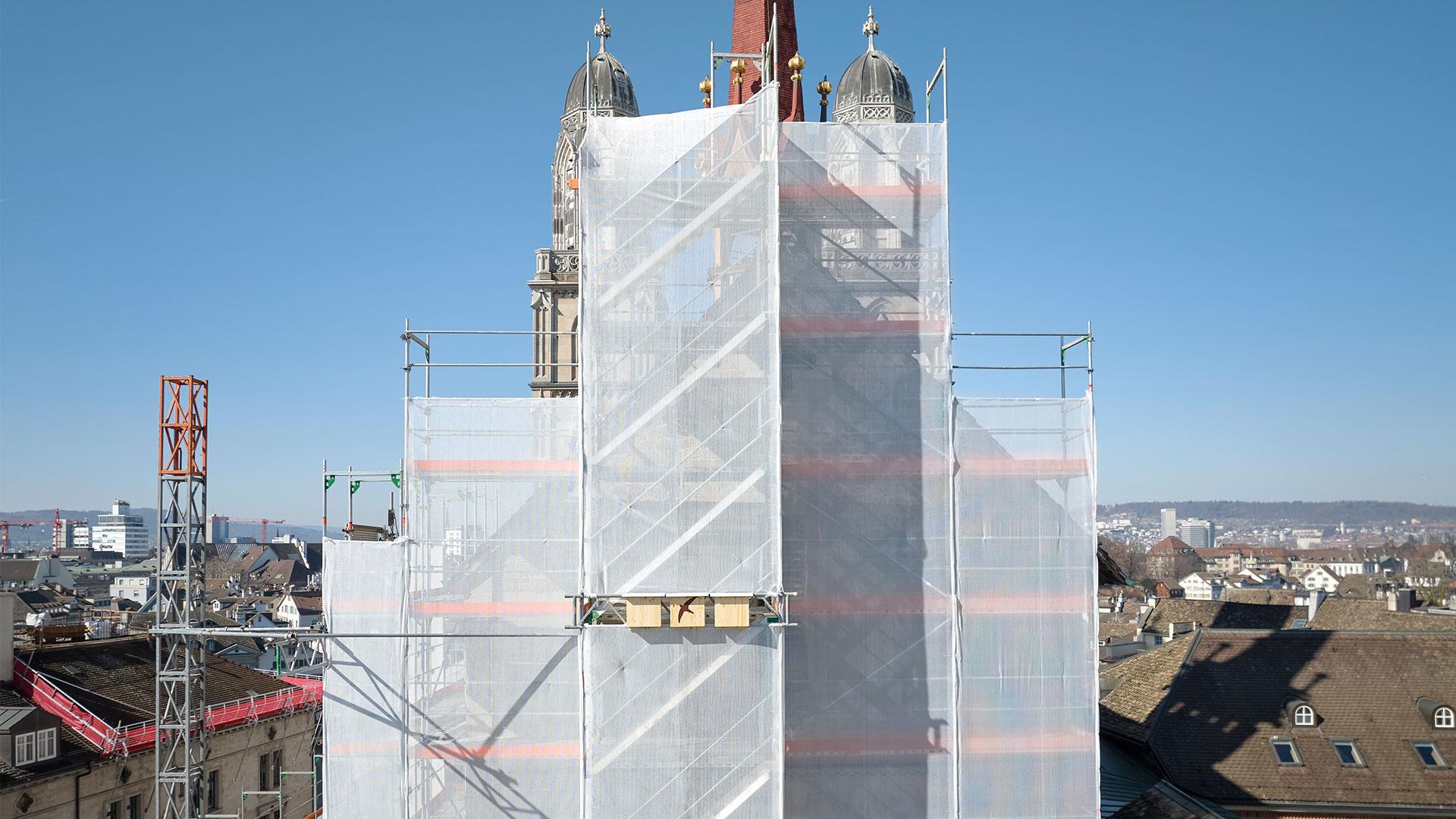Eine hohe Aufnahme eines teilweise mit Baugerüst und weißer Plane verdeckten Kirchturms in Zürich vor blauem Himmel und einer Stadtkulisse.