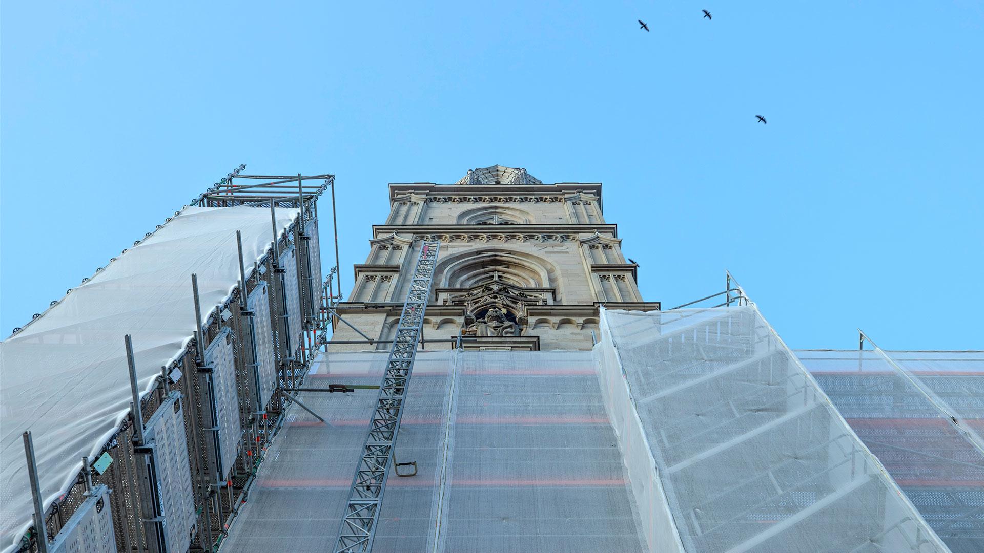 Eine Aufnahme von unten eines Kirchturms in Zürich, teilweise eingerüstet und mit Planen verdeckt, unter einem blauen Himmel mit Vögeln.
