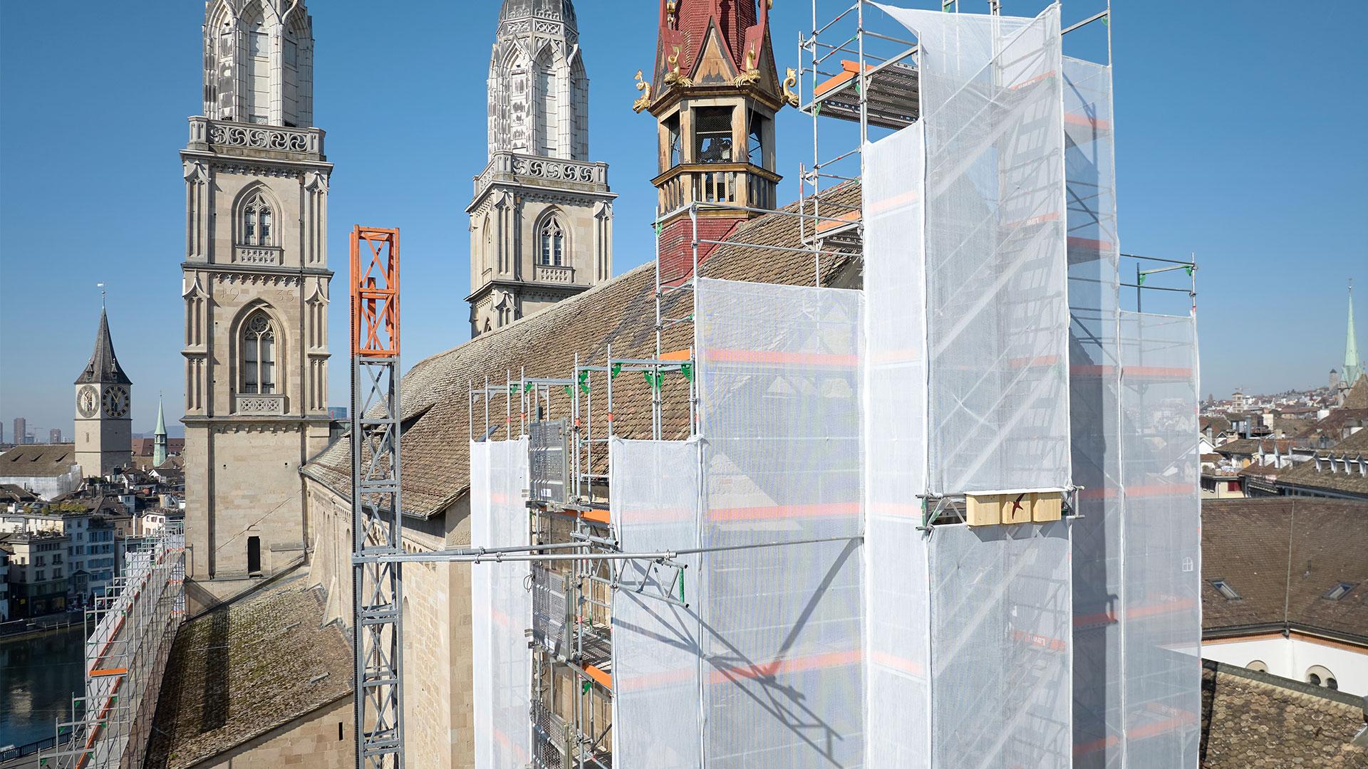 Das Foto zeigt einen Teil des Grossmünsters, teilweise von einem Gerüst mit weissem Netz umgeben. Die markanten Doppeltürme des romanischen Bauwerks ragen links und in der Mitte des Bildes auf. Im Hintergrund ist ein Blick auf die Stadt Zürich mit weiteren Kirchtürmen und Gebäuden unter einem klaren, blauen Himmel zu sehen.